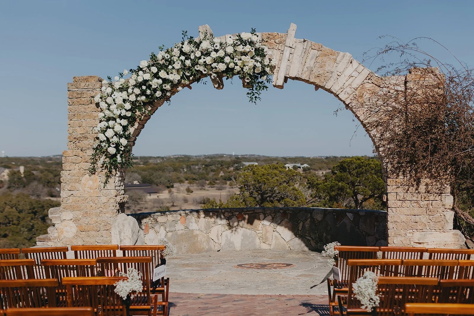 An outdoor wedding altar featuring a stone arch decorated with white flowers, situated in a scenic landscape with trees and open sky, surrounded by wooden chairs.
