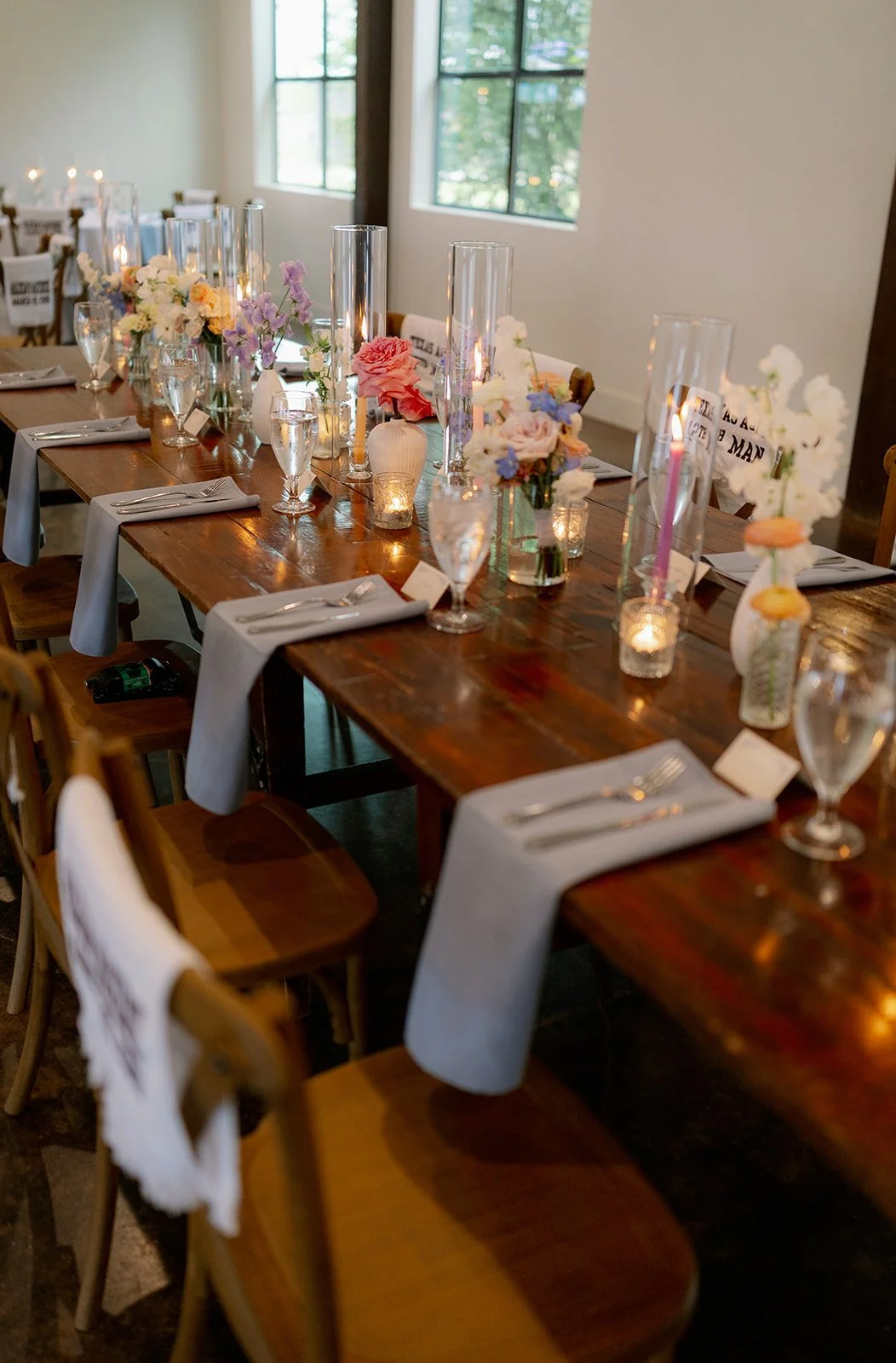 A decorated wooden dining table set for a formal event with white napkins, silverware, wine glasses, and floral centerpieces in small vases. Candleholders with candles are also on the table, with natural light coming through large windows in the background.