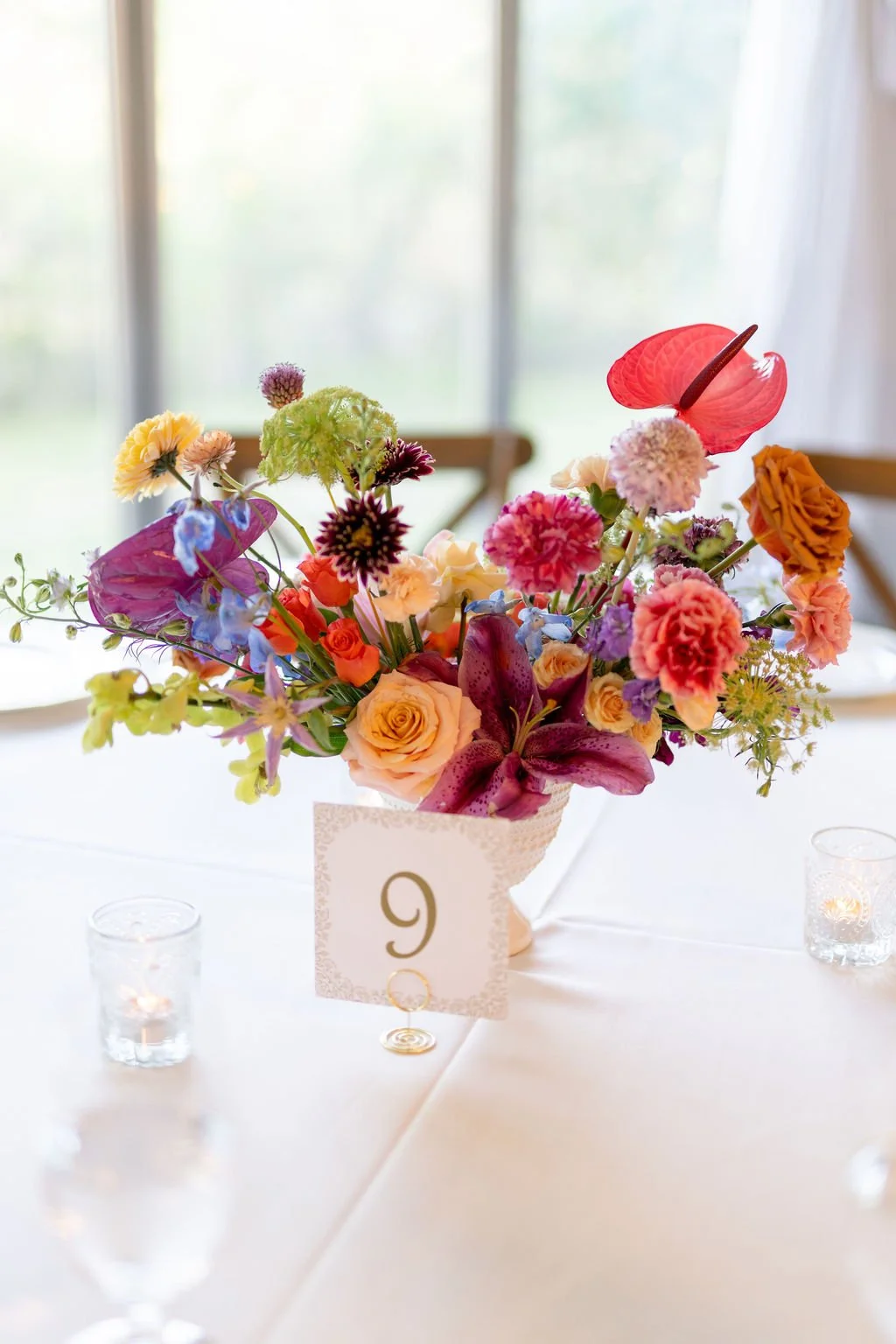 A colorful floral centerpiece with various flowers including lilies, roses, carnations, and anthuriums in a white vase on a decorated table with a table number '9' and small candles.