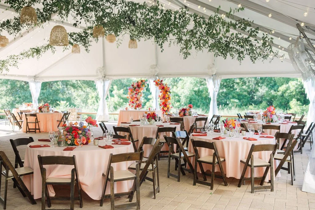 Decorated outdoor party tent with pink tablecloth-covered tables, floral centerpieces, and string lights with greenery overhead.
