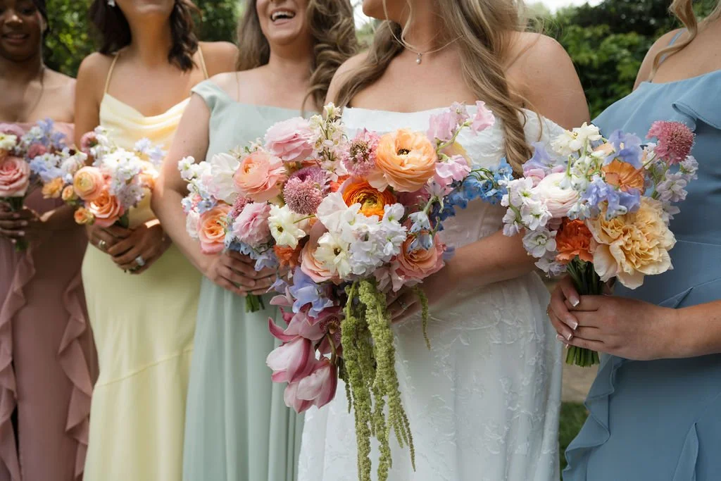 A group of women wearing pastel dresses holding colorful bouquets of flowers at an outdoor wedding.