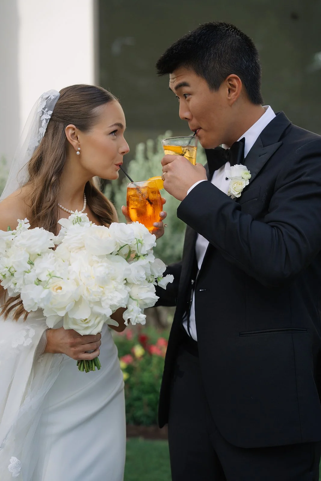 Bride and groom sharing drinks at their wedding, with bride holding a bouquet of white flowers and wearing a wedding dress and veil, groom in a tuxedo with a boutonniere, outdoors with a blurred natural background.