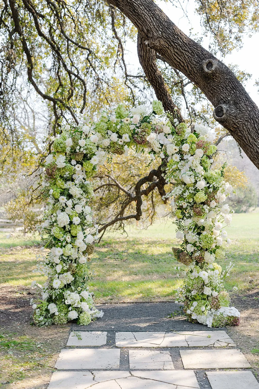 A floral wedding arch made of white and green flowers stands under a large tree in an outdoor setting.