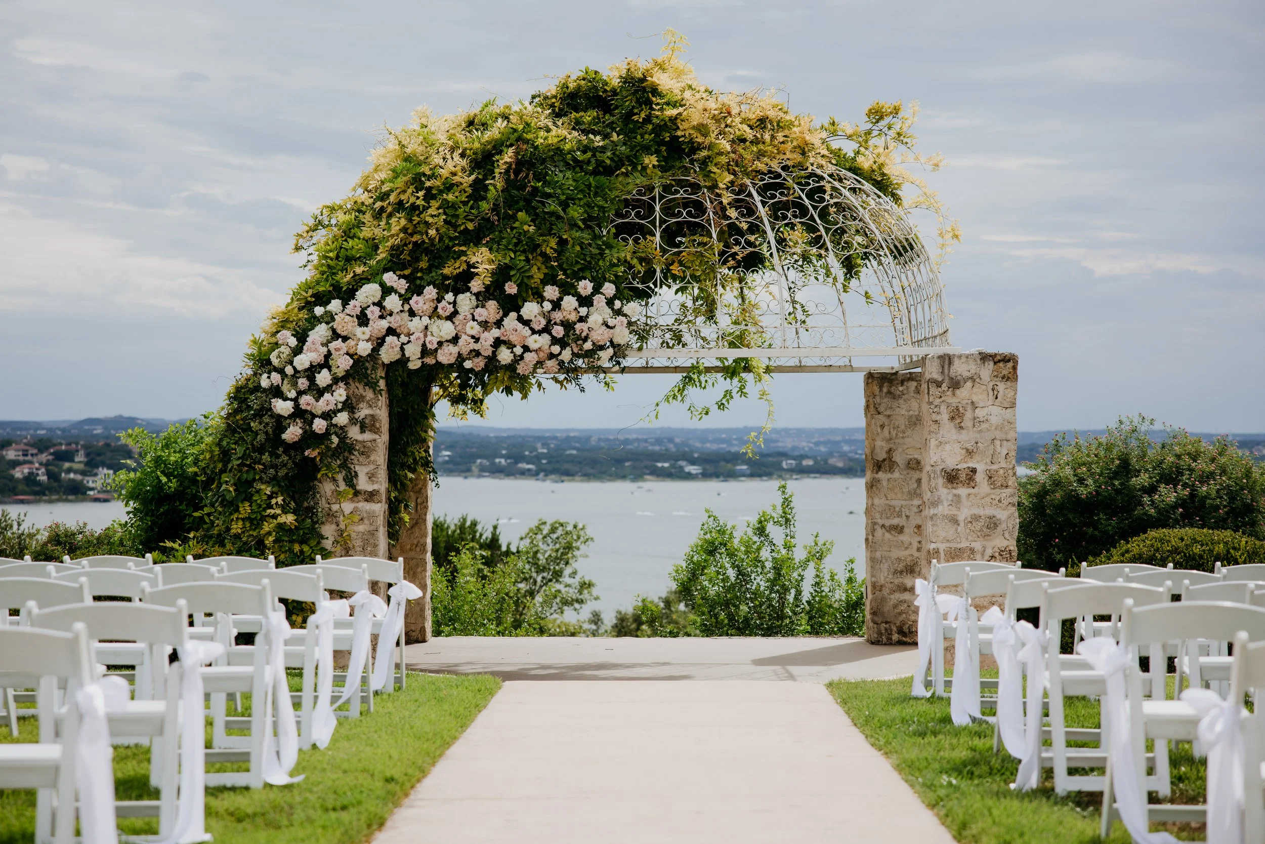 Outdoor wedding ceremony setup with white chairs, floral archway, and scenic water view.