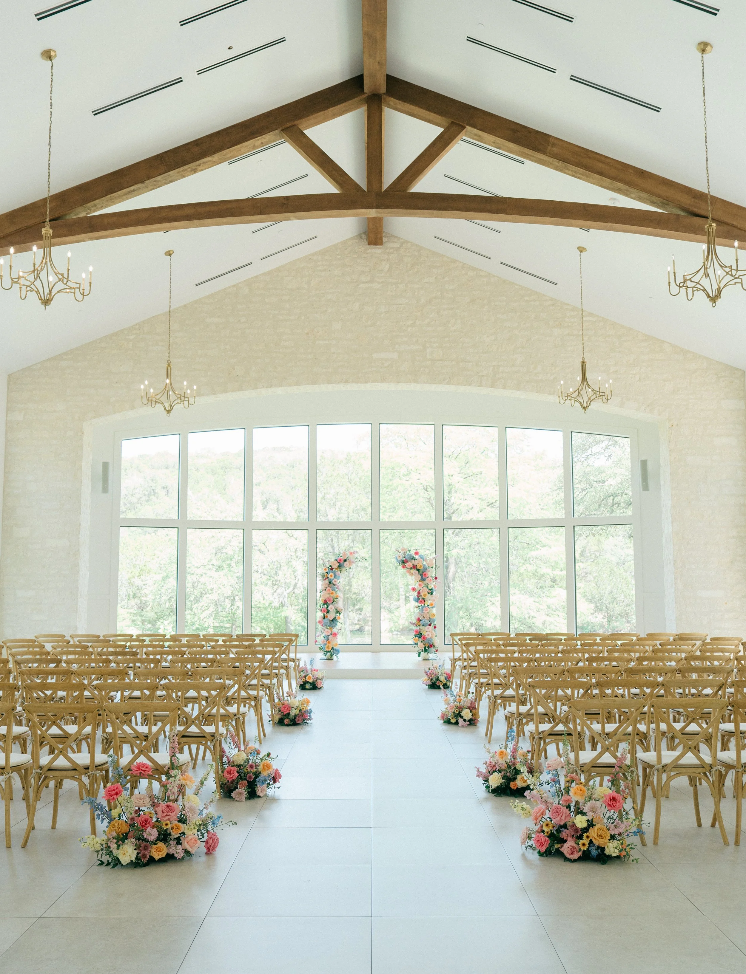 Wedding ceremony setup inside a bright room with large windows, wooden chairs arranged in rows, floral decorations along the aisle, and floral arch at the front.