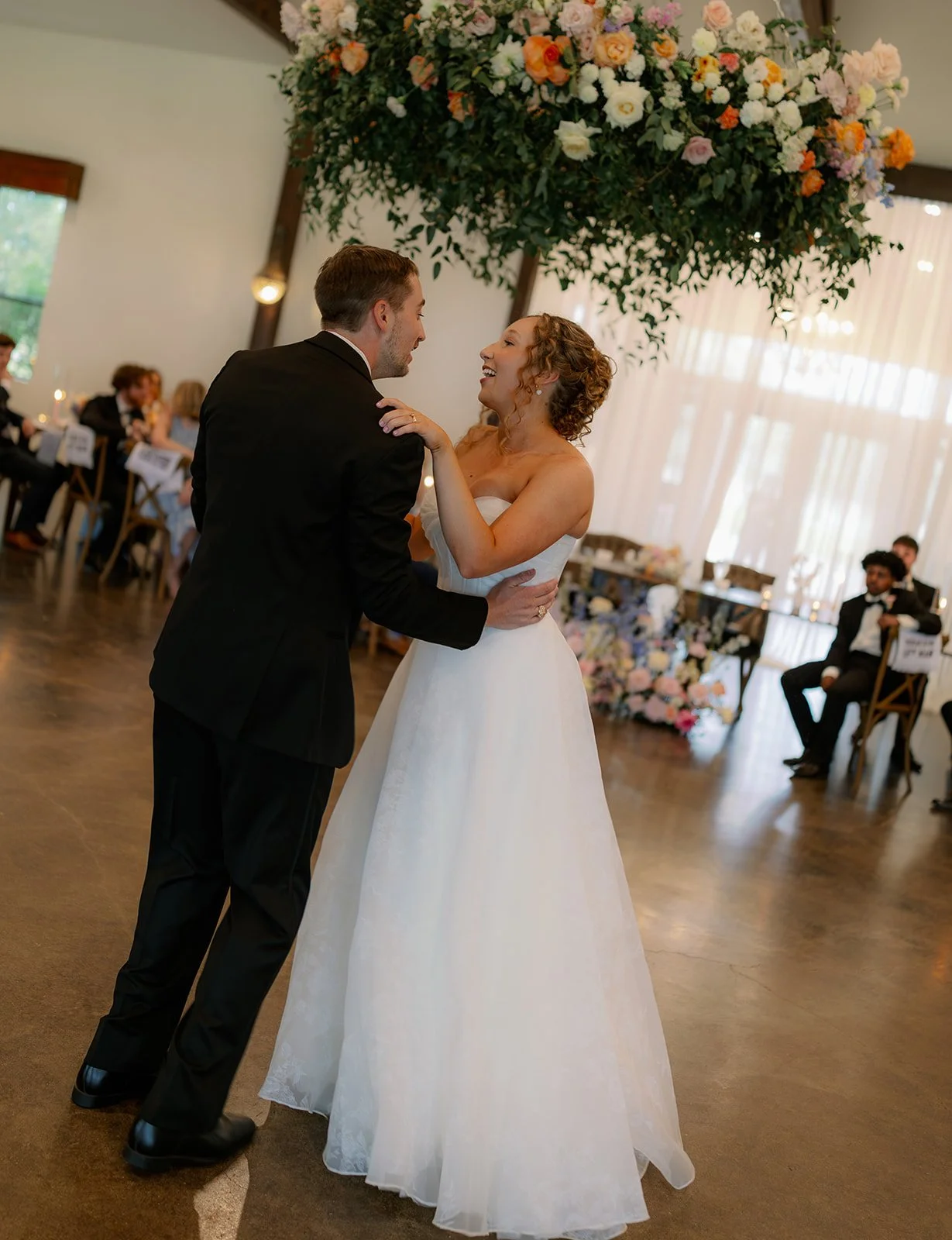 A bride and groom dancing at their wedding reception, with guests seated at decorated tables in the background.