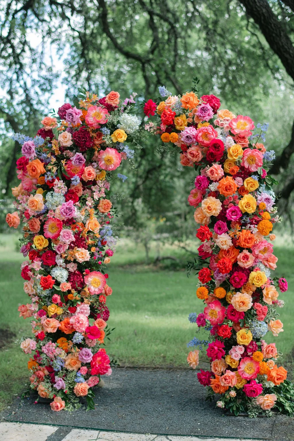 Colorful floral arch made of pink, red, orange, yellow, purple, and white flowers set outdoors with green trees in the background.