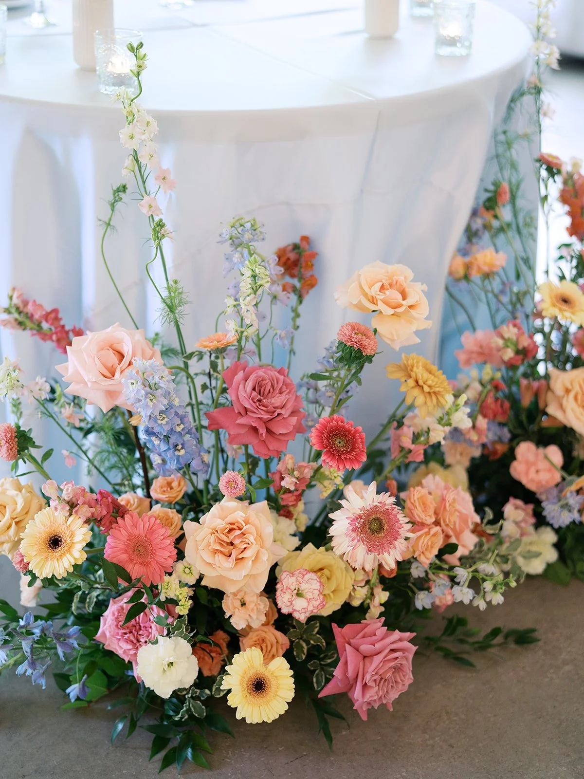 Colorful floral arrangement with roses, daisies, and other flowers in soft pastel shades on a beige carpet in front of a white table draped with a white tablecloth.