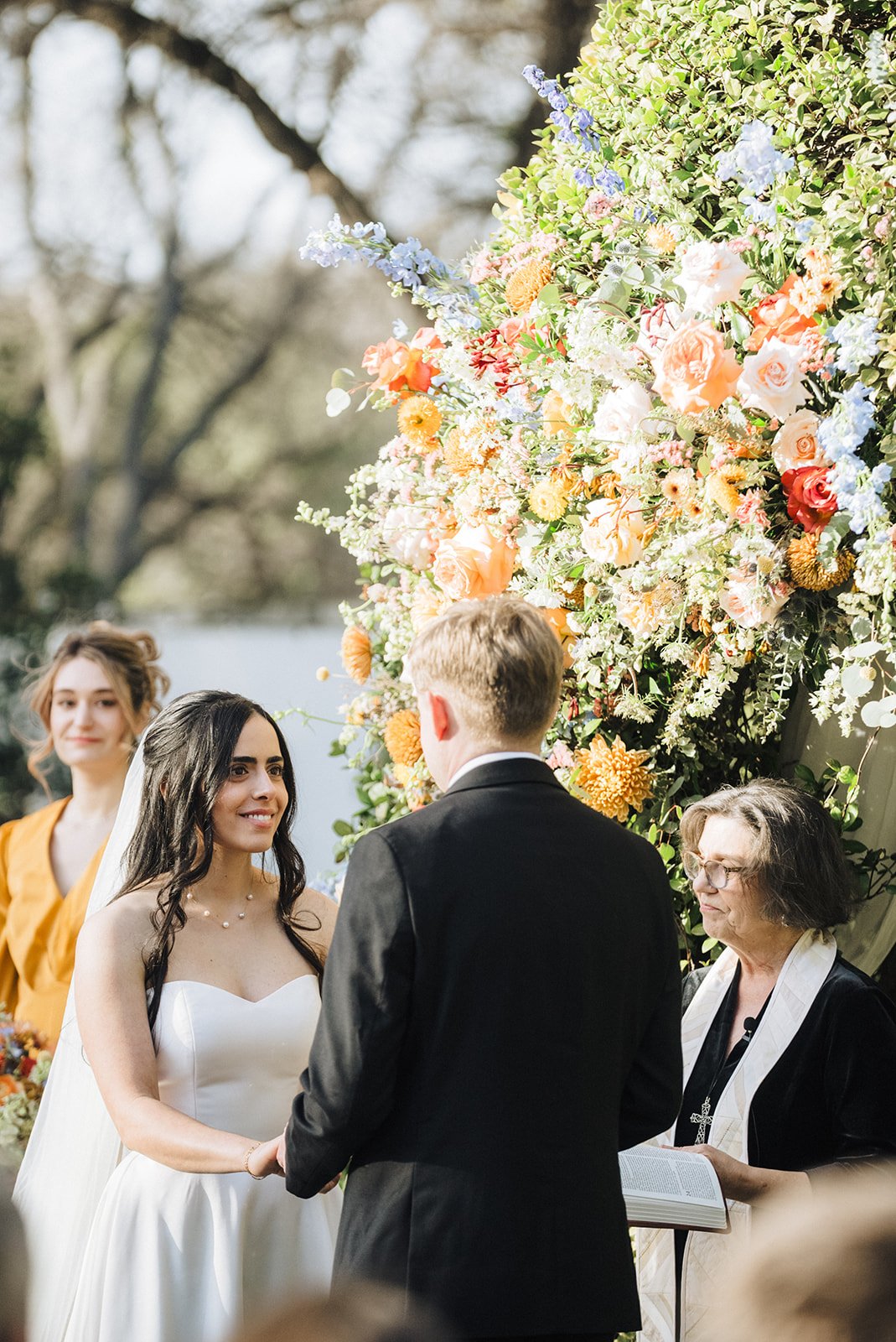 A wedding ceremony outdoors with a couple exchanging vows in front of a large floral backdrop, including roses and other colorful flowers, with an officiant and a bridesmaid present.