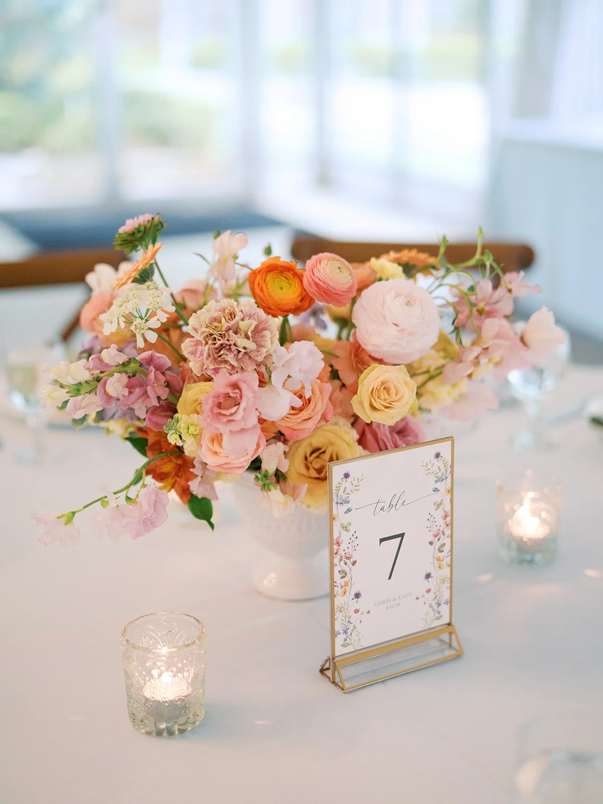 A floral centerpiece with pink, peach, and yellow flowers on a white table, with a table number card for table 7 and small candle holders with lit candles surrounding it, at a bright indoor event.
