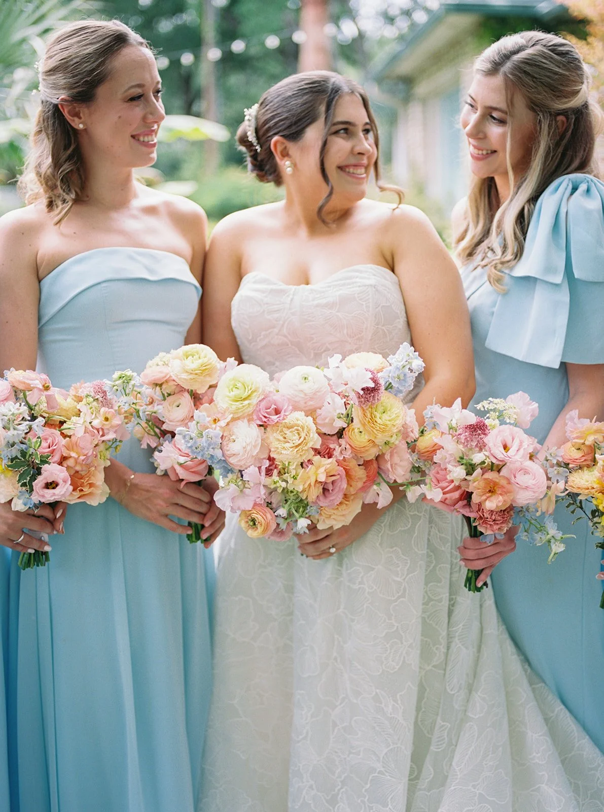 Three women in pastel dresses holding bouquets of pink, purple, and cream flowers at a garden wedding.
