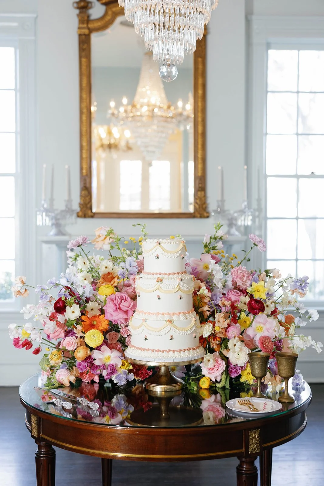 A three-tiered cake decorated with pink and white icing, surrounded by colorful flowers and placed on a dark wooden table with two gold goblets and a plate with a fork. The setting features a large mirror, chandeliers, and bright windows.