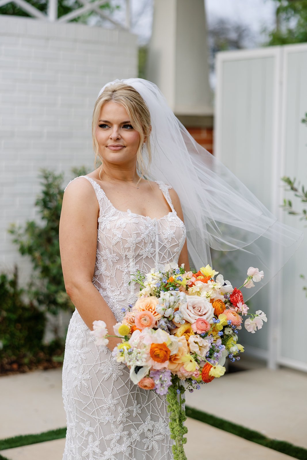 A bride in a white lace wedding dress holding a colorful bouquet of flowers, standing outdoors with greenery and a white fence in the background.