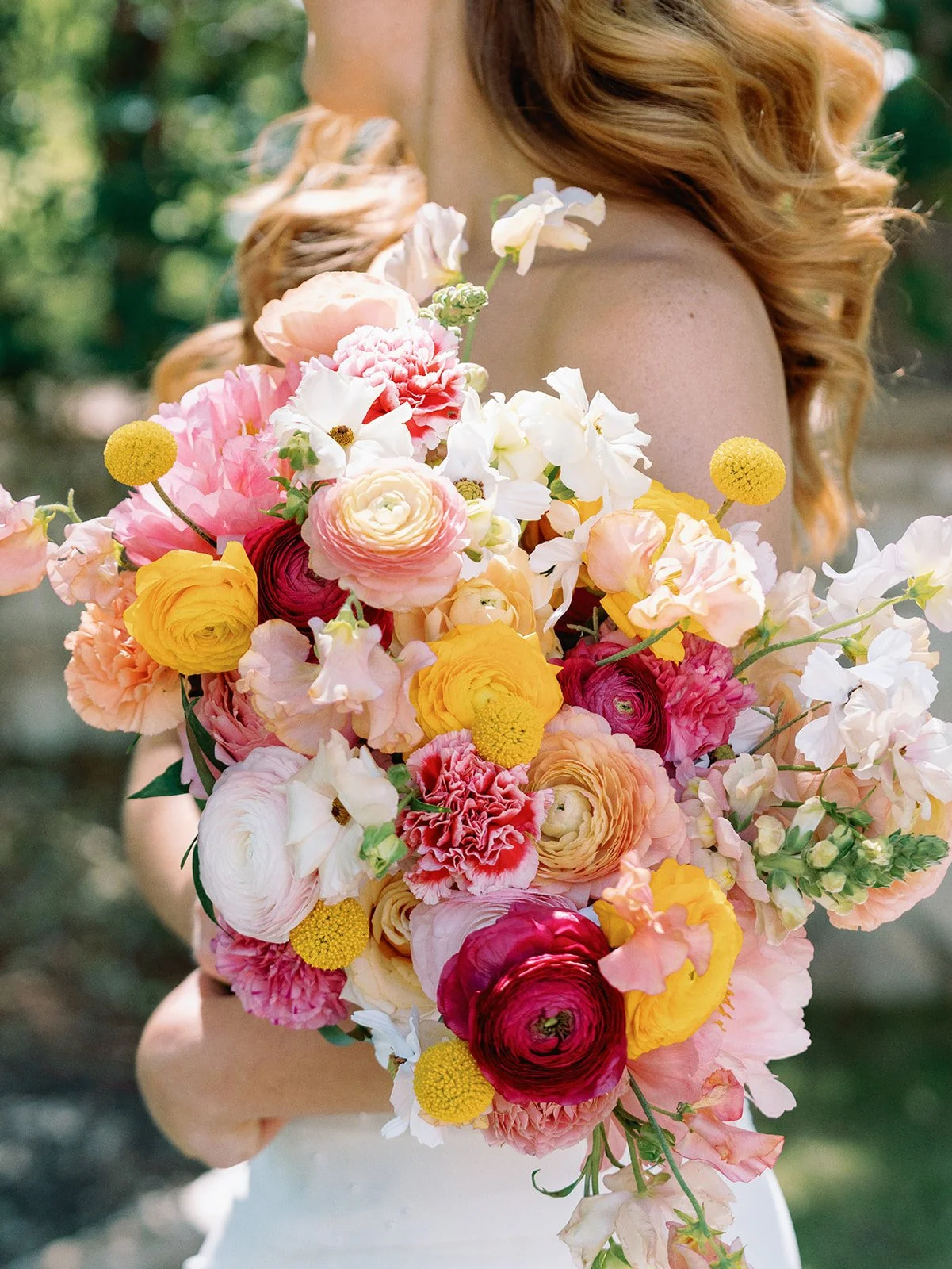 A woman with curly red hair holding a large, colorful bouquet of various flowers including roses, ranunculus, and daisies outdoors.