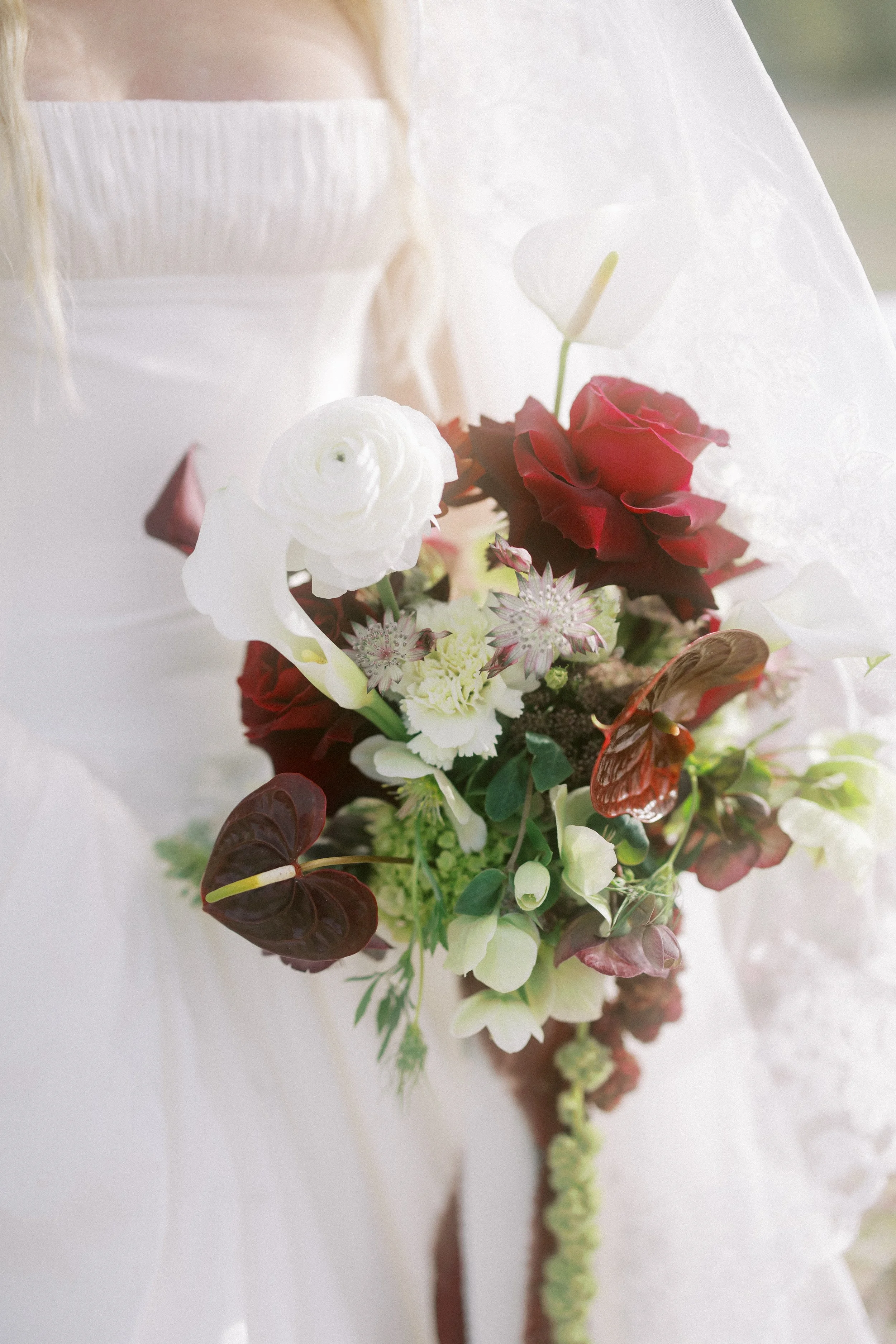 A bridal bouquet with white, red, and pink flowers, including roses, orchids, and other blooms, held against white fabric.