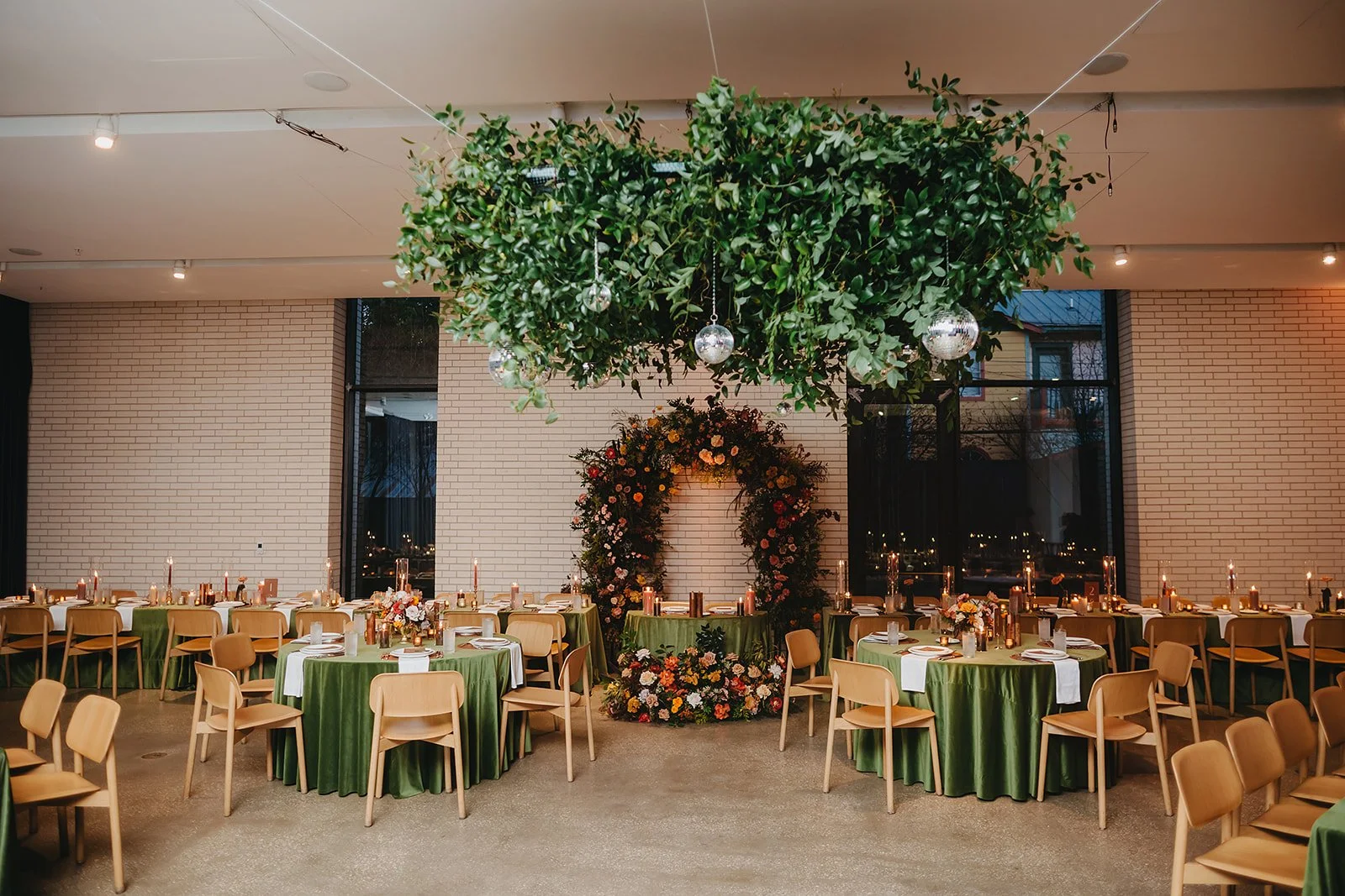 Elegantly decorated banquet hall with round tables covered in green tablecloths, floral centerpieces, and candles. A large greenery installation with hanging disco balls decorates the ceiling, and a floral arch is centered at the back of the room. Th