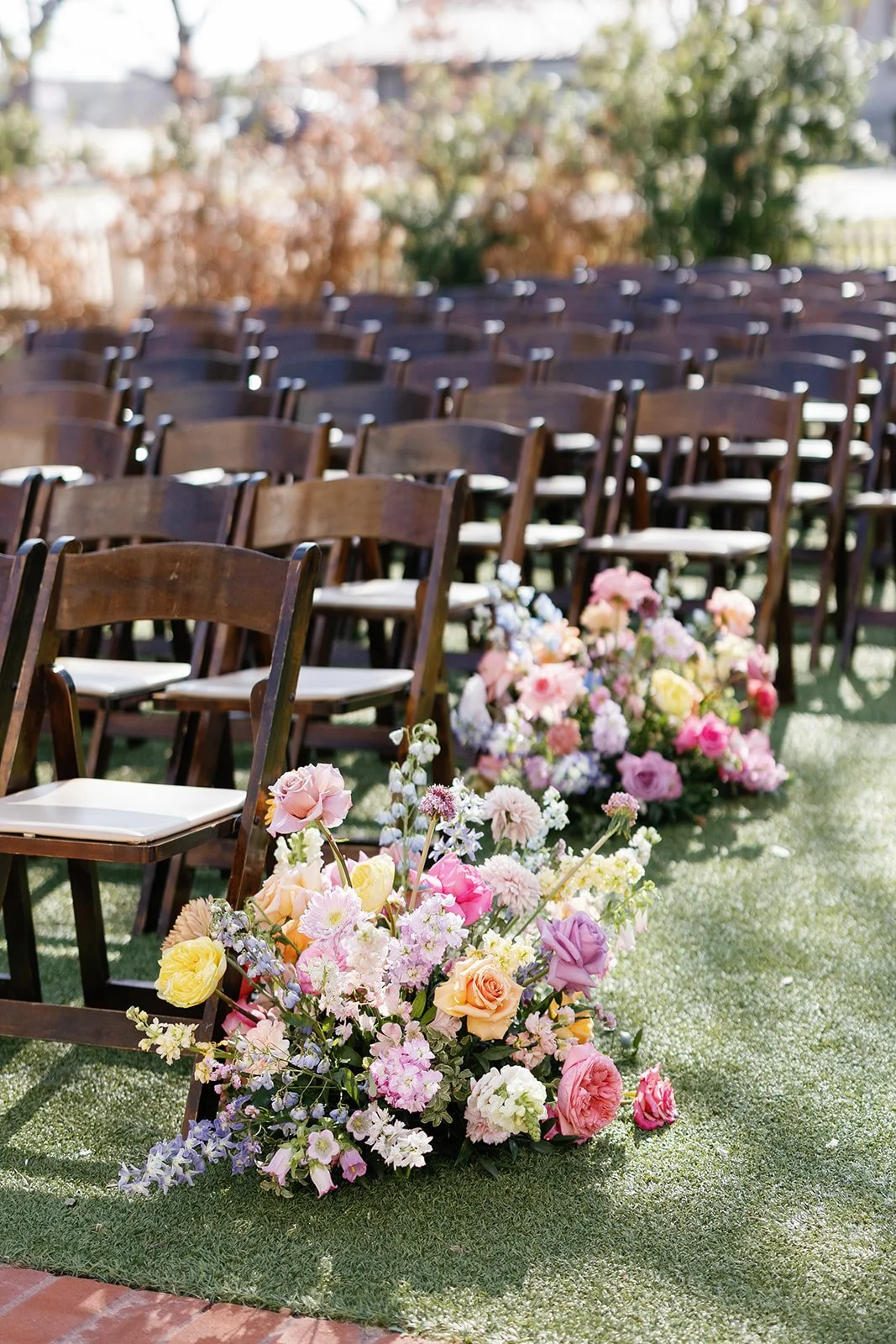 Rows of wooden chairs at an outdoor wedding ceremony decorated with a large arrangement of pink, yellow, and white flowers on the grass.