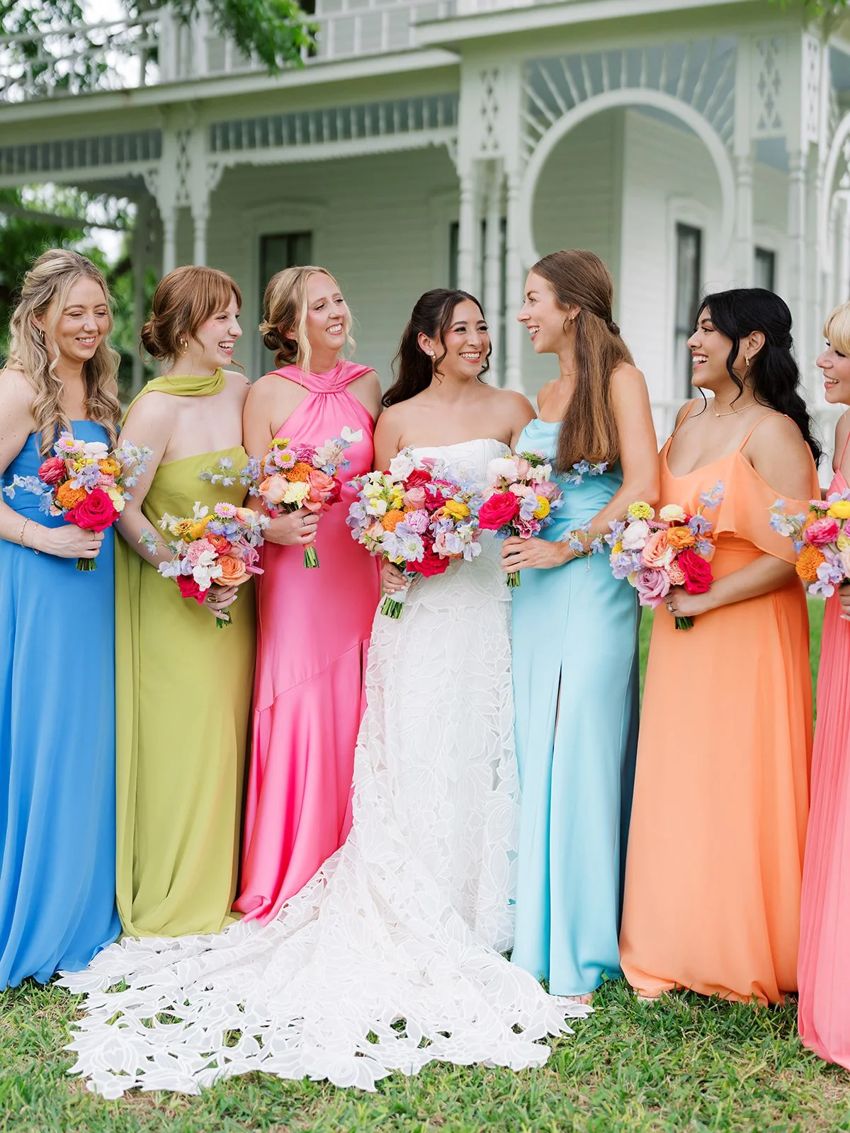 Group of women in colorful dresses holding bouquets of flowers, standing outdoors in front of a house, celebrating a wedding.