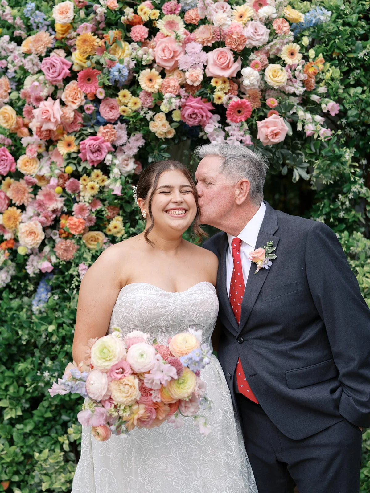 An elderly man in a dark suit with a red polka dot tie kisses a young woman in a white wedding dress on the cheek, standing in front of a colorful flower wall, holding a bouquet of pastel flowers.