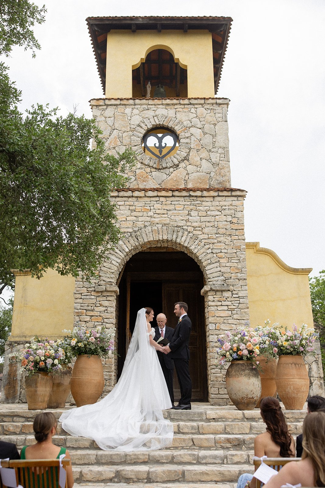 A wedding ceremony taking place outdoors in front of a stone and stucco building with a clock tower, decorated with large flower arrangements in clay pots, with a bride and groom exchanging vows.