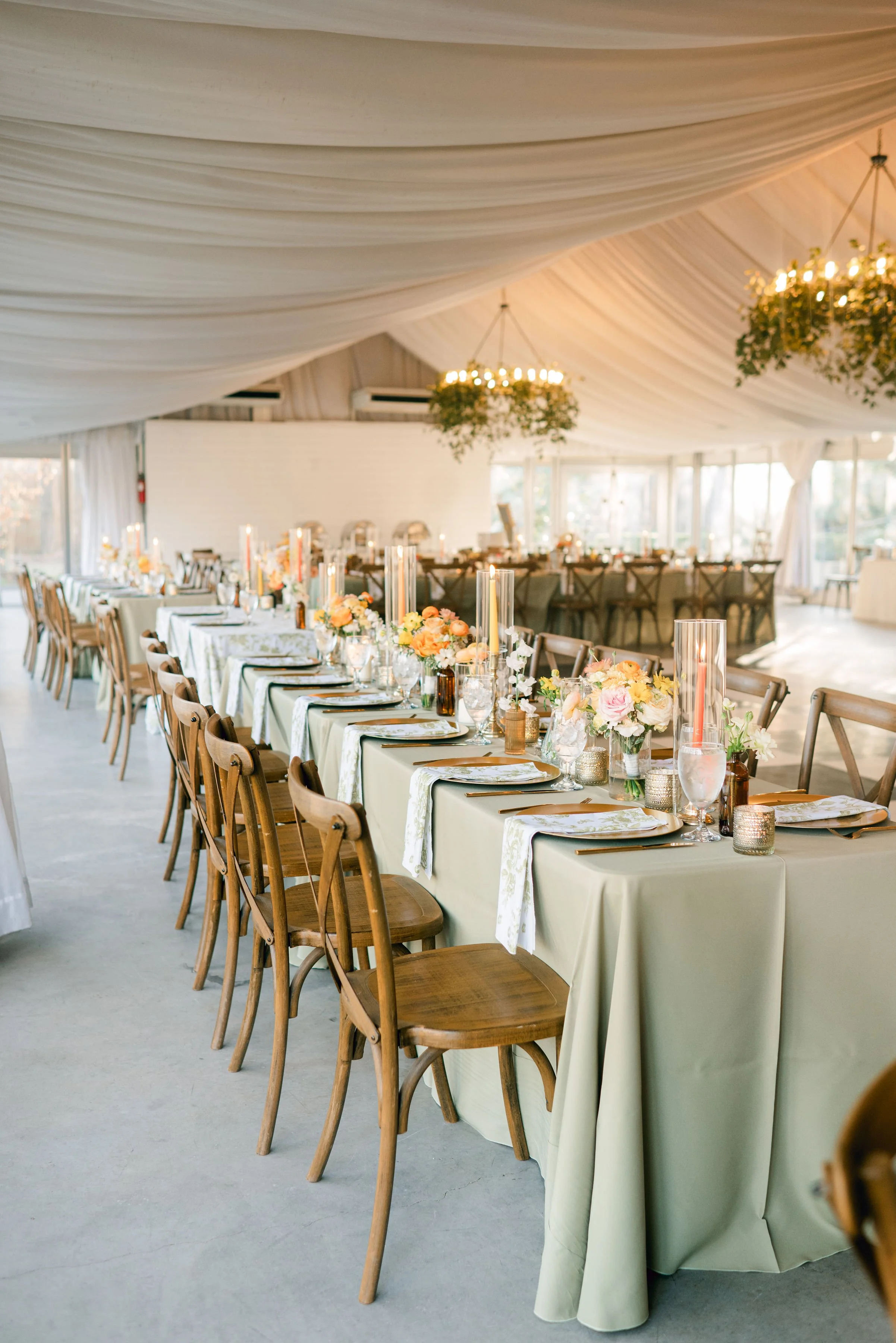 Indoor wedding reception with long table decorated with flowers, candles, and place settings, under a ceiling draped with white fabric and two chandeliers with greenery.