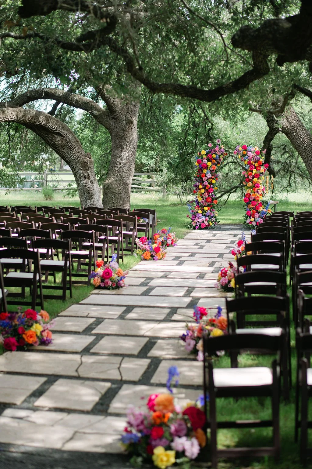 An outdoor wedding setup with rows of black chairs on a stone path, decorated with colorful flower arrangements, leading to a floral arch under large trees in a green park.