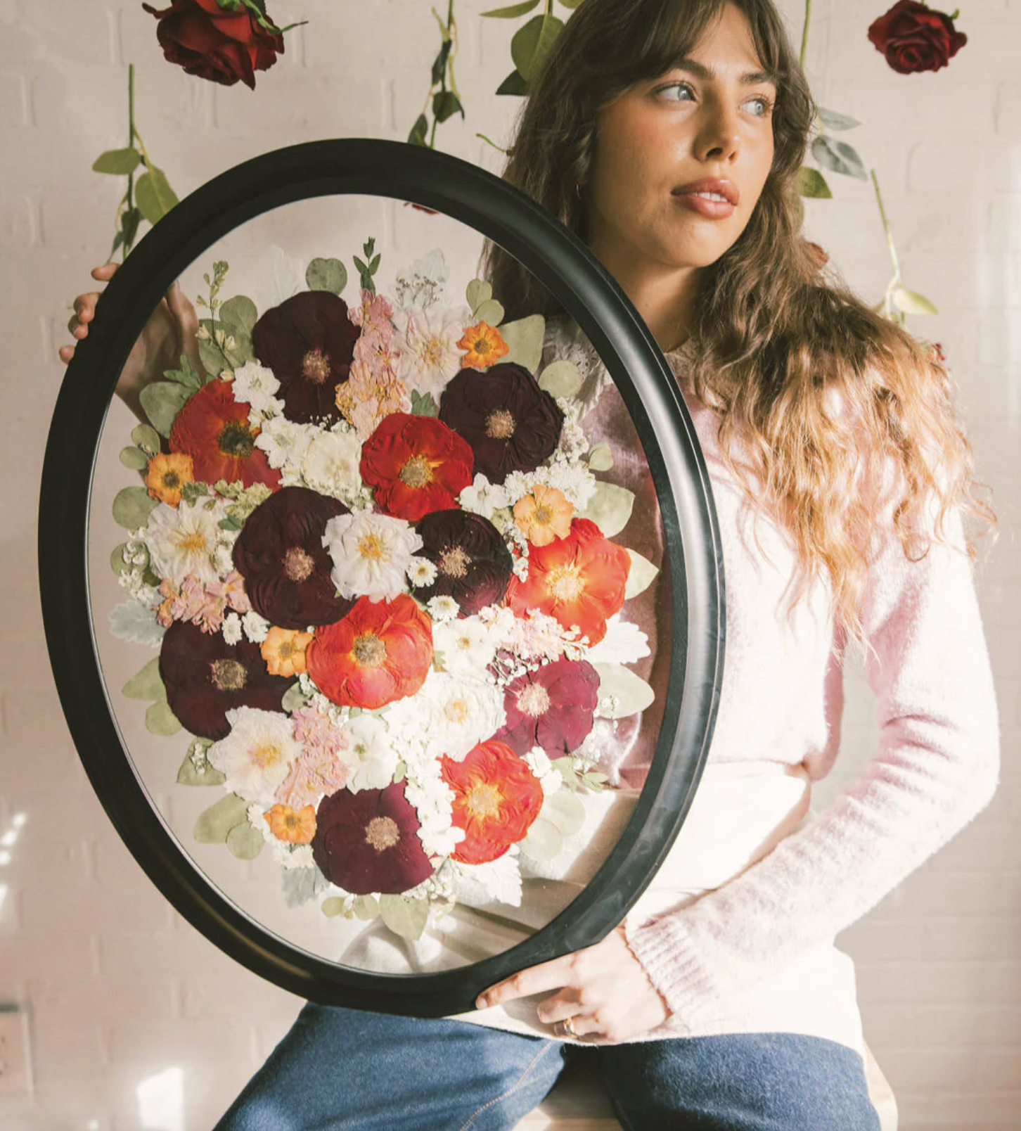 A woman with wavy brown hair wearing a pink sweater and blue jeans holding a large black oval frame with a floral arrangement inside. The arrangement includes red, white, pink, and orange flowers with green leaves, set against a white brick wall background.