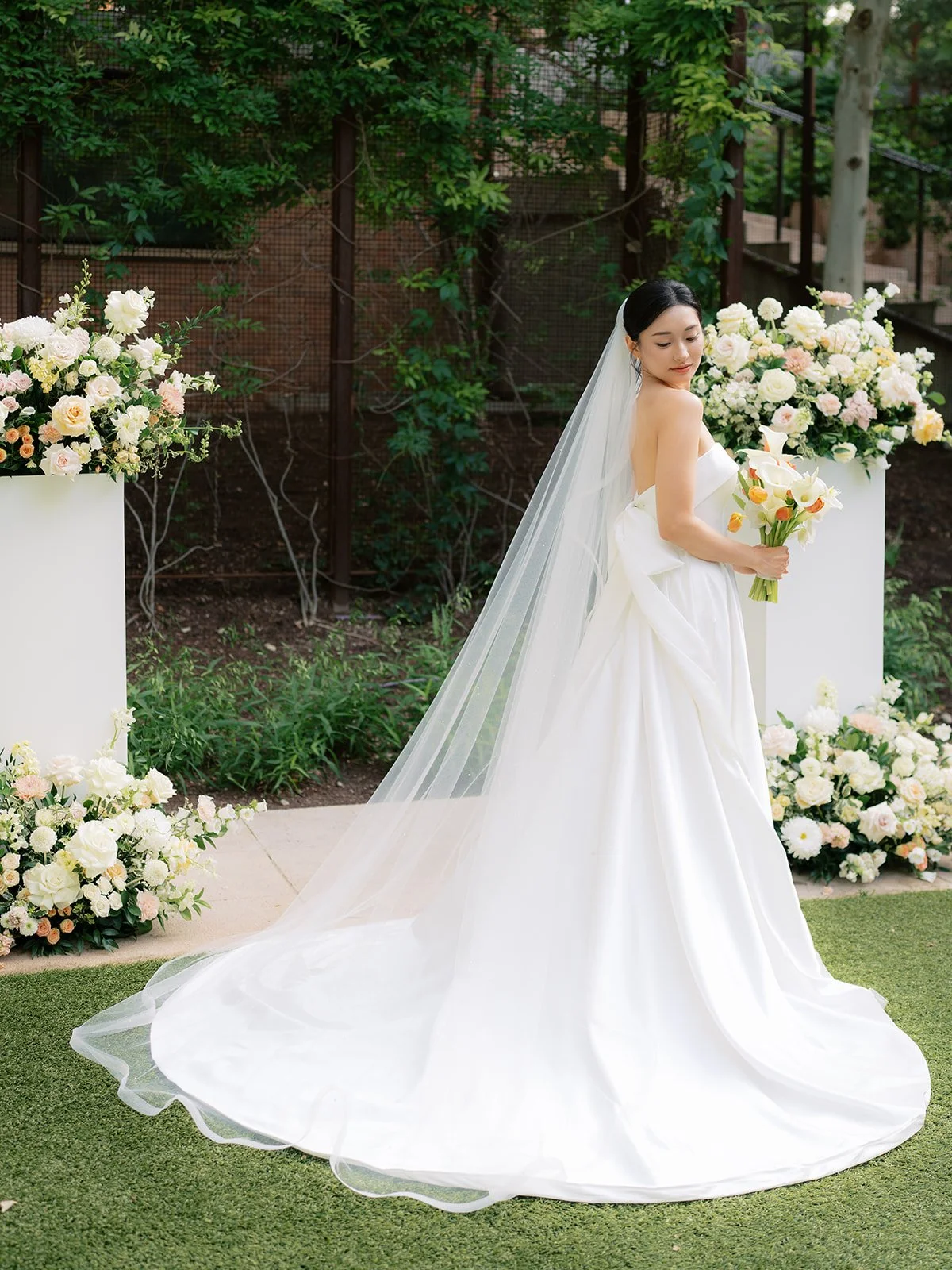 A bride in a white wedding gown with a veil holding a bouquet of flowers, standing outdoors surrounded by floral arrangements and greenery.