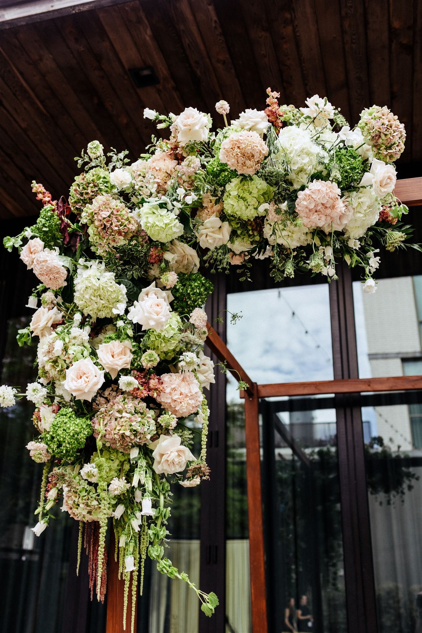 Large floral arrangement with white, pink, and green flowers hanging from a wooden structure indoors.