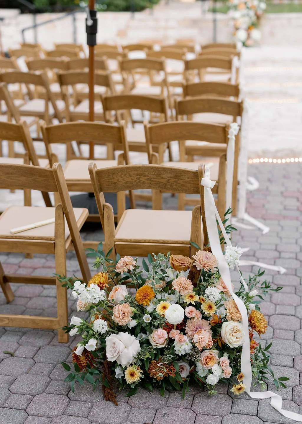 Wedding ceremony setup with wooden chairs and floral arrangement at the front, outdoors on a brick pathway.