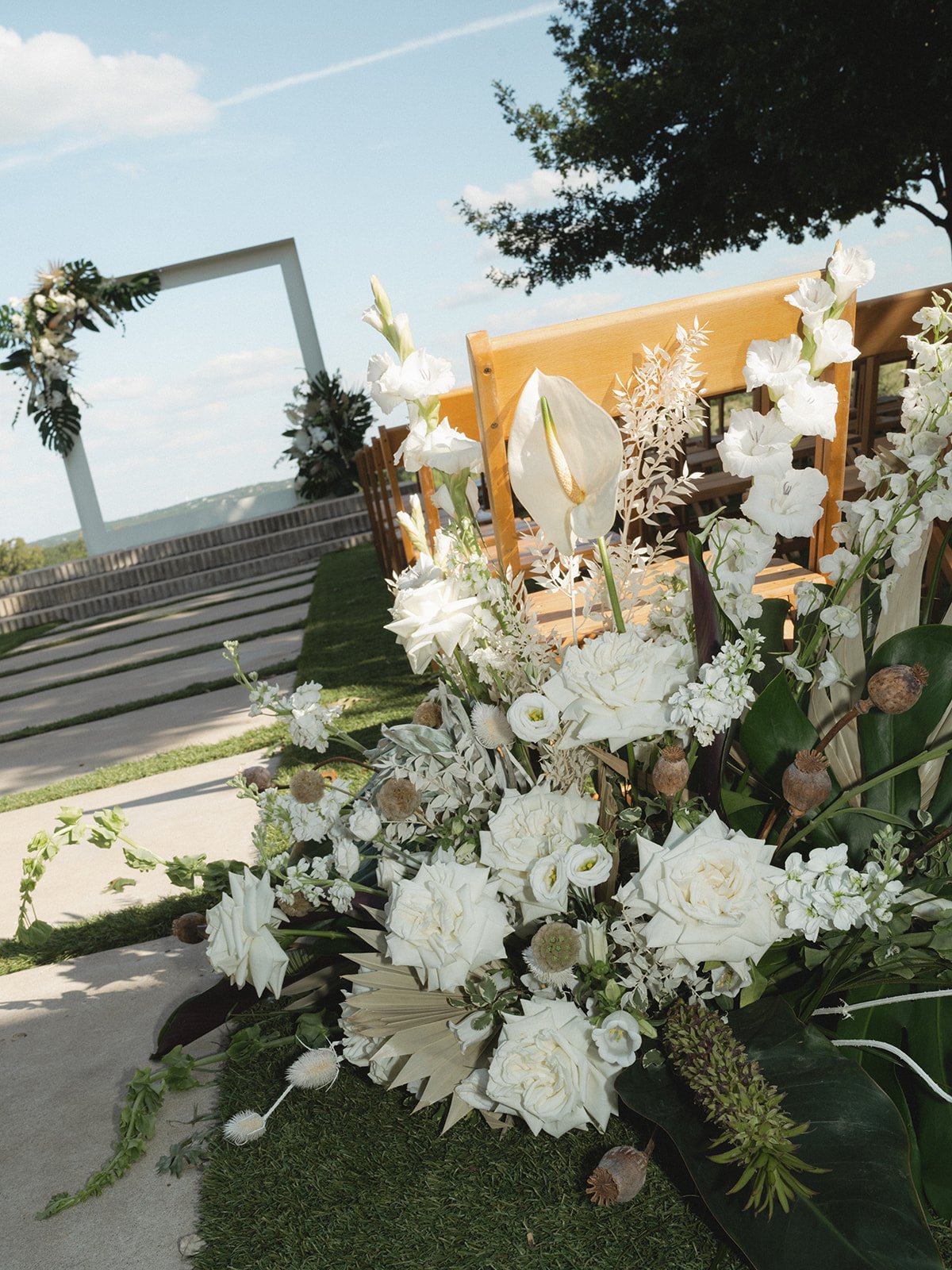 A wedding ceremony setup outdoors with a floral arrangement of mostly white flowers and greenery in the foreground, wooden benches, and a white arch decorated with white flowers and greenery in the background under a blue sky.