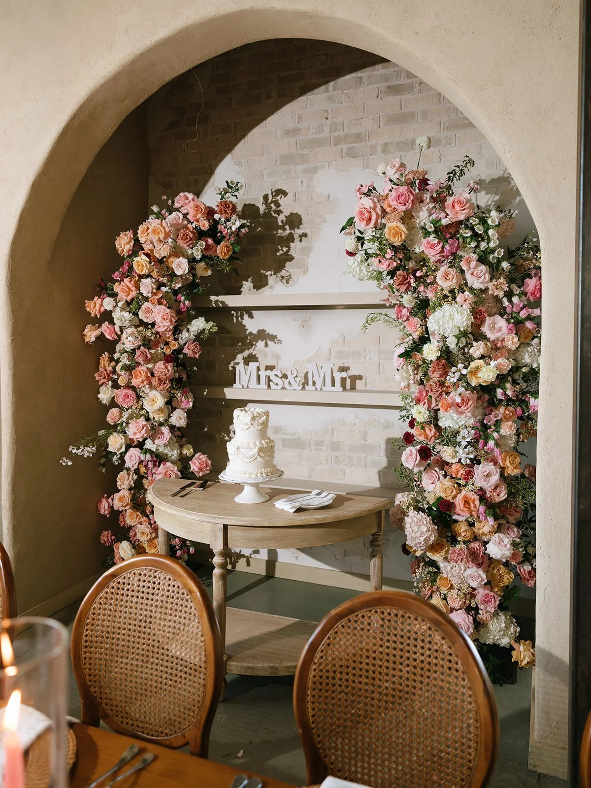 Wedding decor with a floral arch, a white wedding cake on a small round table, and a sign that reads 'Mrs & Mr' against a brick wall, framed by an archway.