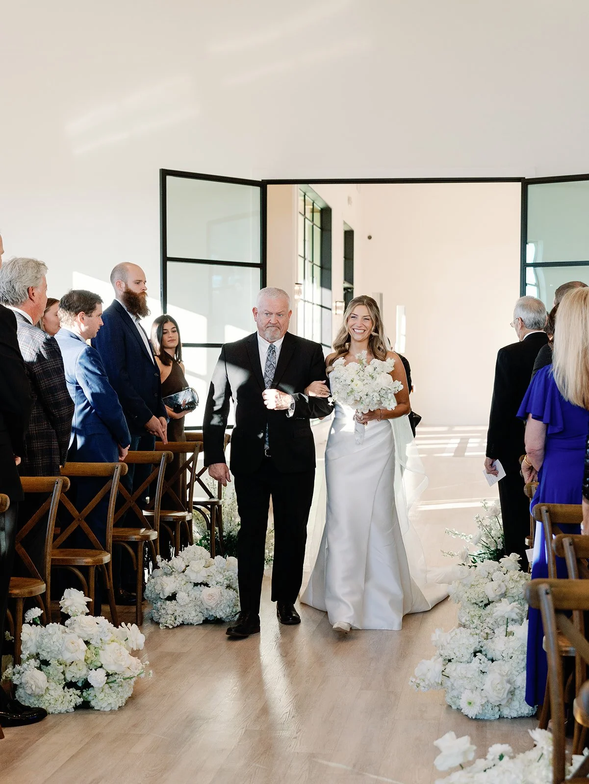 Bride walking down the aisle with her father during a wedding ceremony, surrounded by guests and floral decorations.