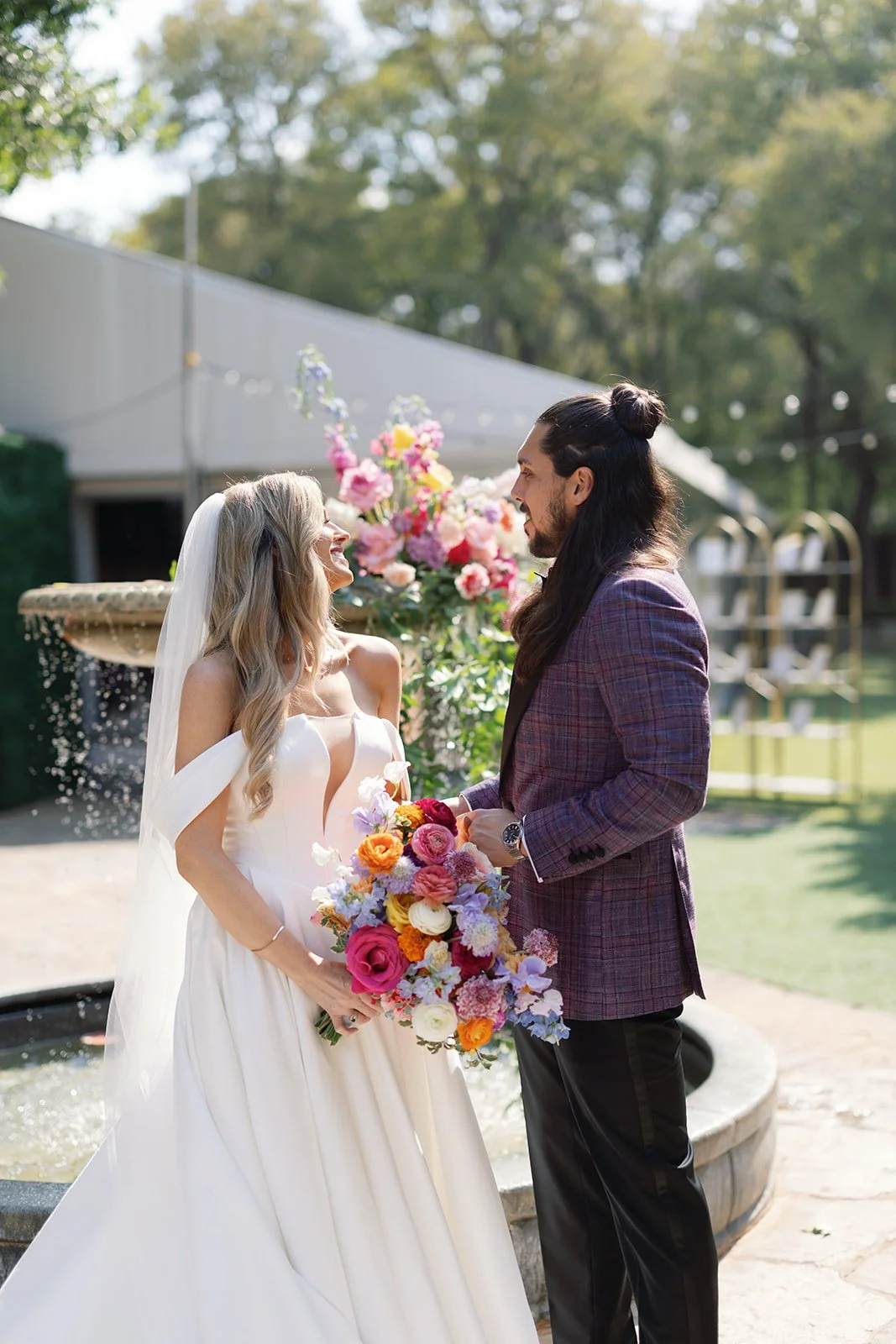 Bride and groom standing outdoors, holding a colorful bouquet, smiling at each other during their wedding ceremony.