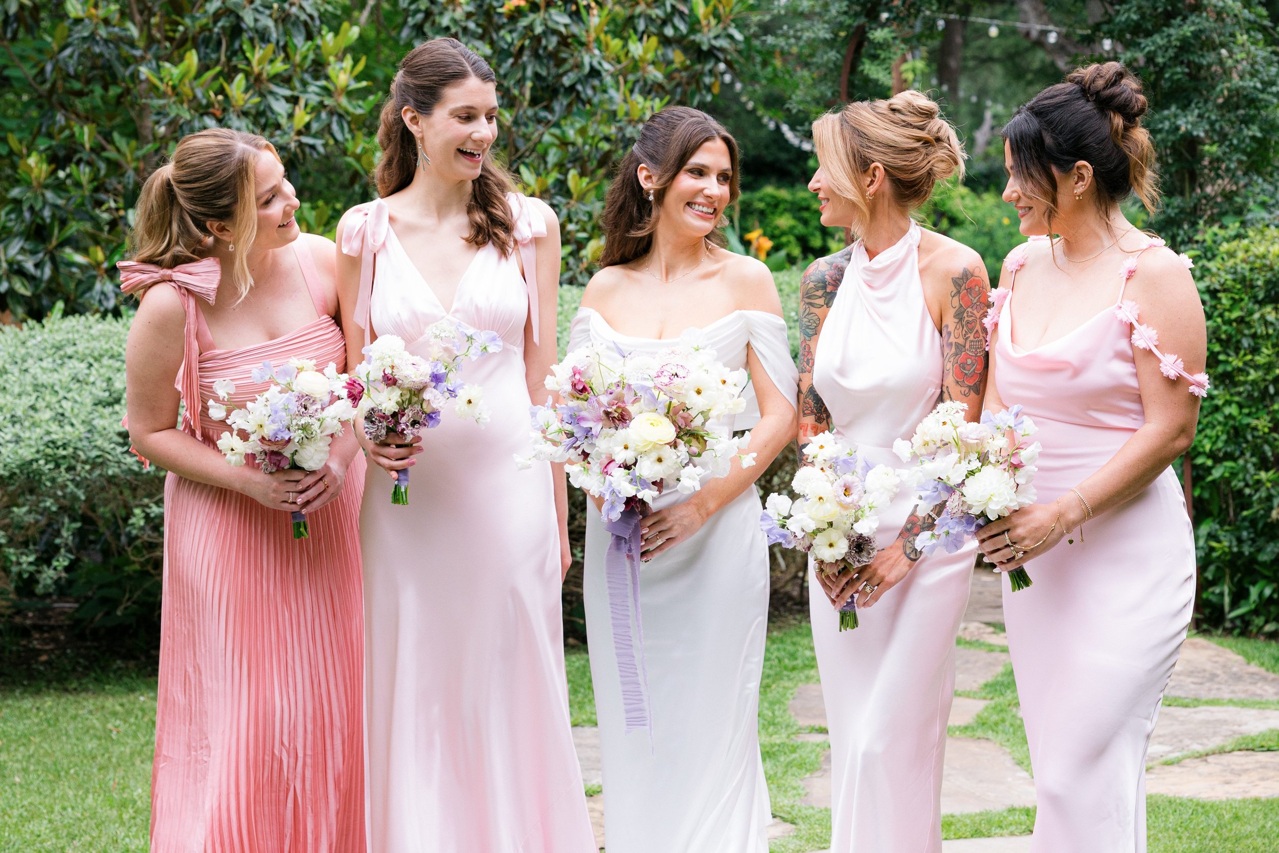 Group of six women smiling and holding bouquets of flowers at an outdoor wedding, standing on a lawn with trees and string lights in the background.