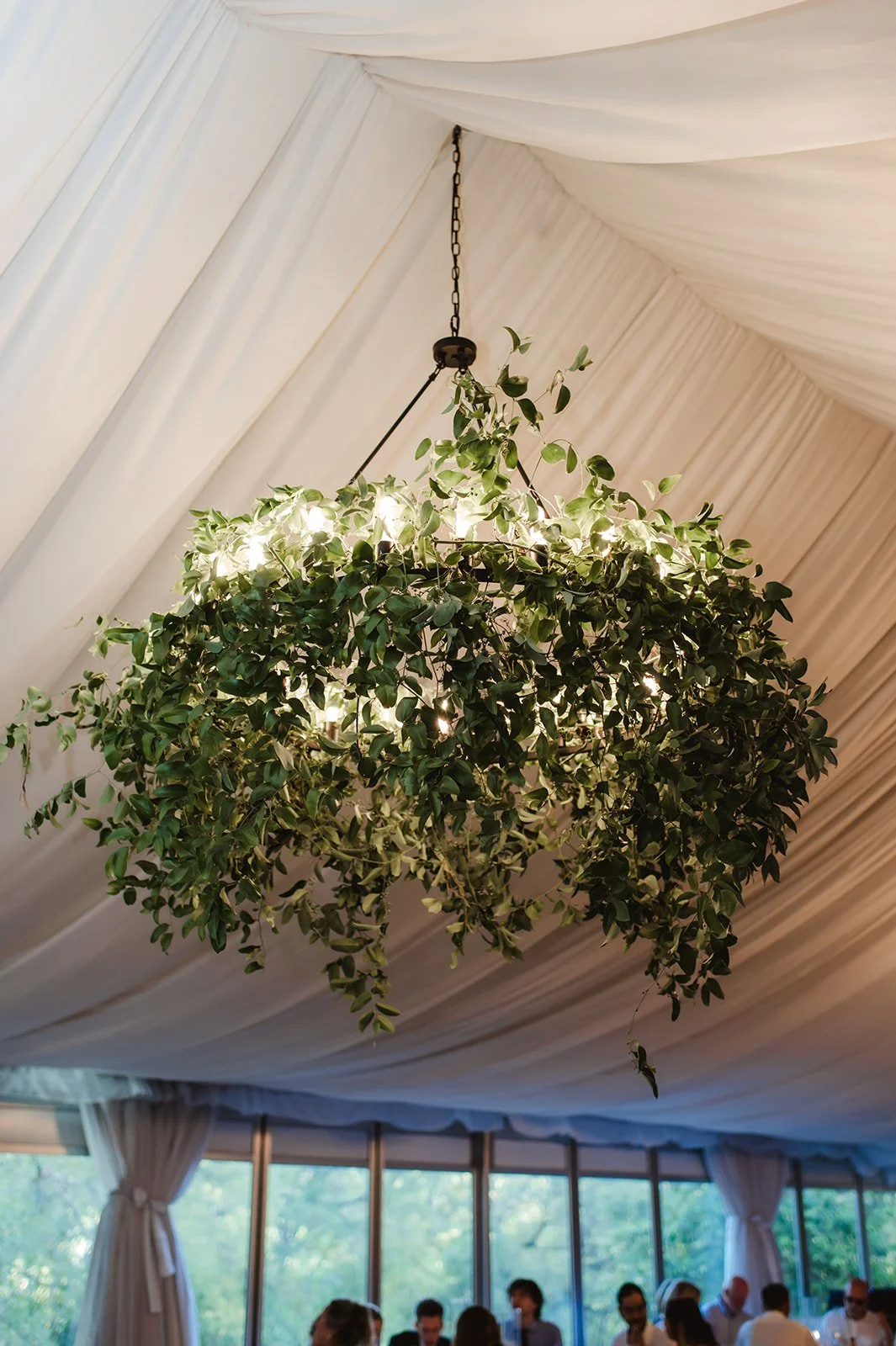 A hanging light fixture designed as a spherical arrangement of green leaves, illuminated from within, hanging from a ceiling with white drapery at an event with people seated at tables.