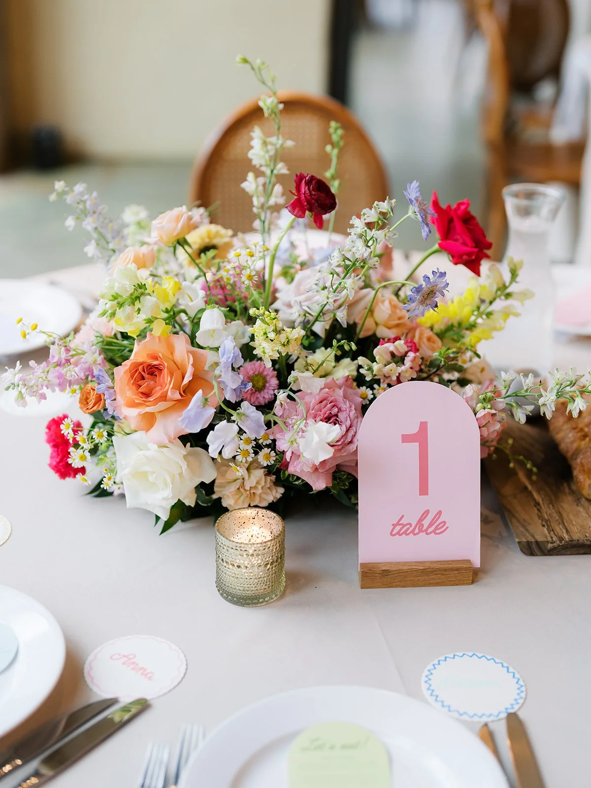 A floral centerpiece on a table with a pink table number sign, a candle, and place cards. The flowers are in pastel and bright colors, including roses, daisies, and snapdragons.