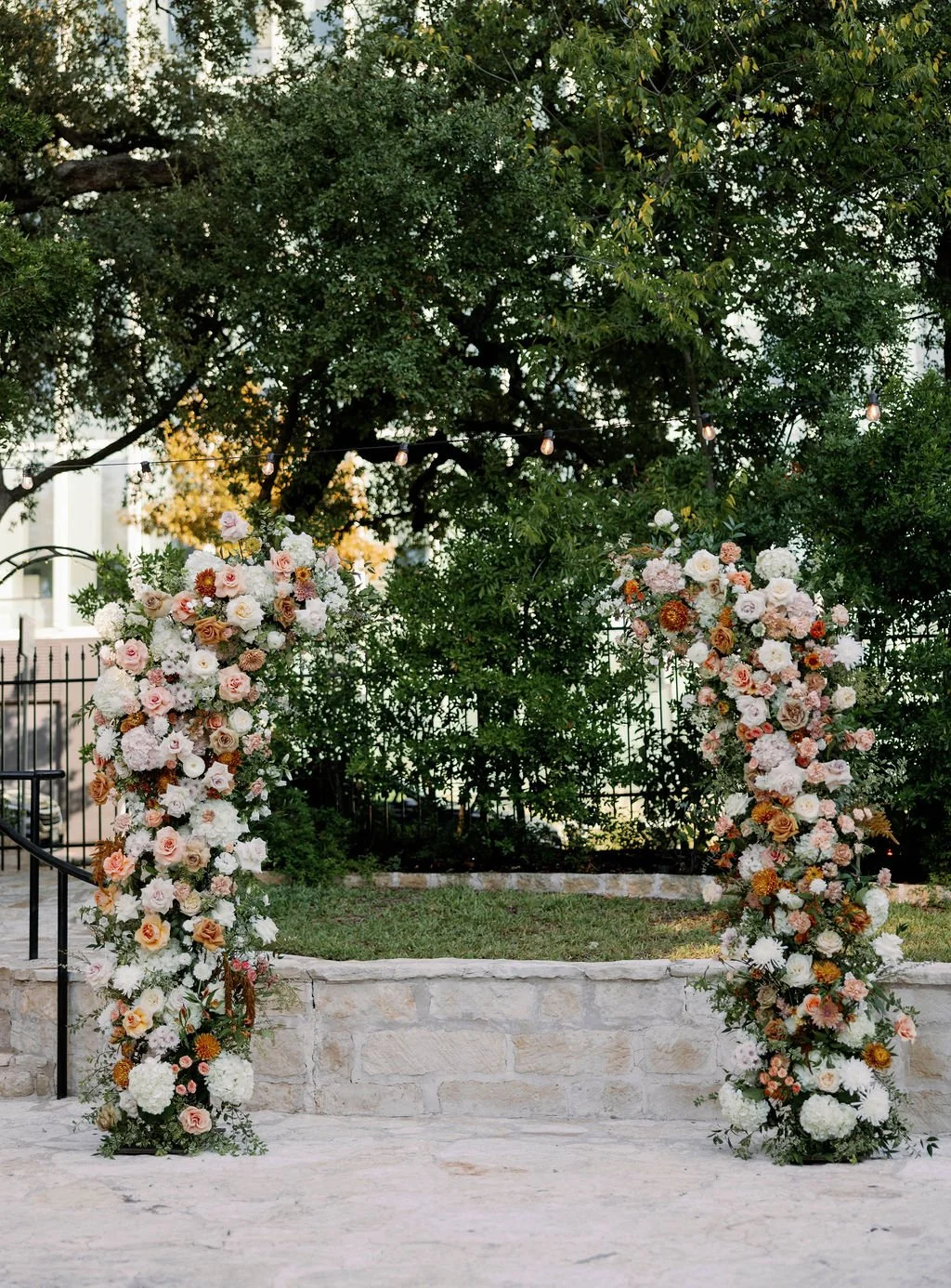 A floral wedding arch decorated with white, pink, and peach roses and other flowers, set outdoors with greenery and trees in the background.