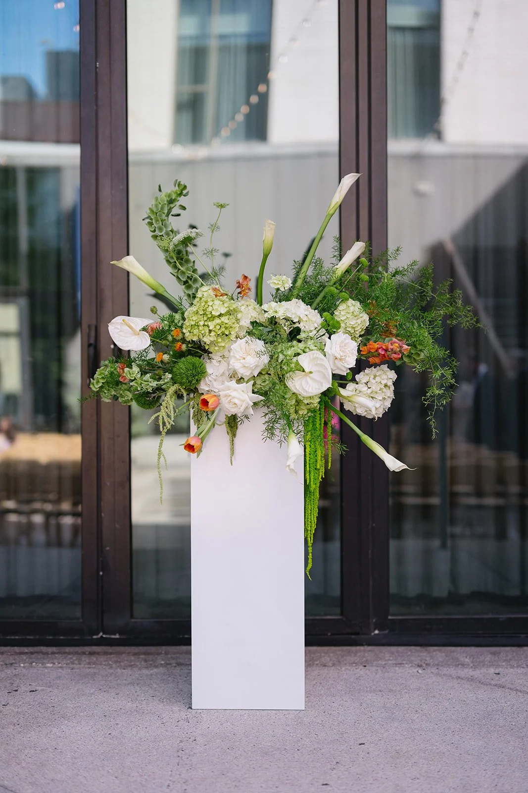 A tall white vase with a large arrangement of white and green flowers, including calla lilies, hydrangeas, and other greenery, placed outside in front of glass doors.