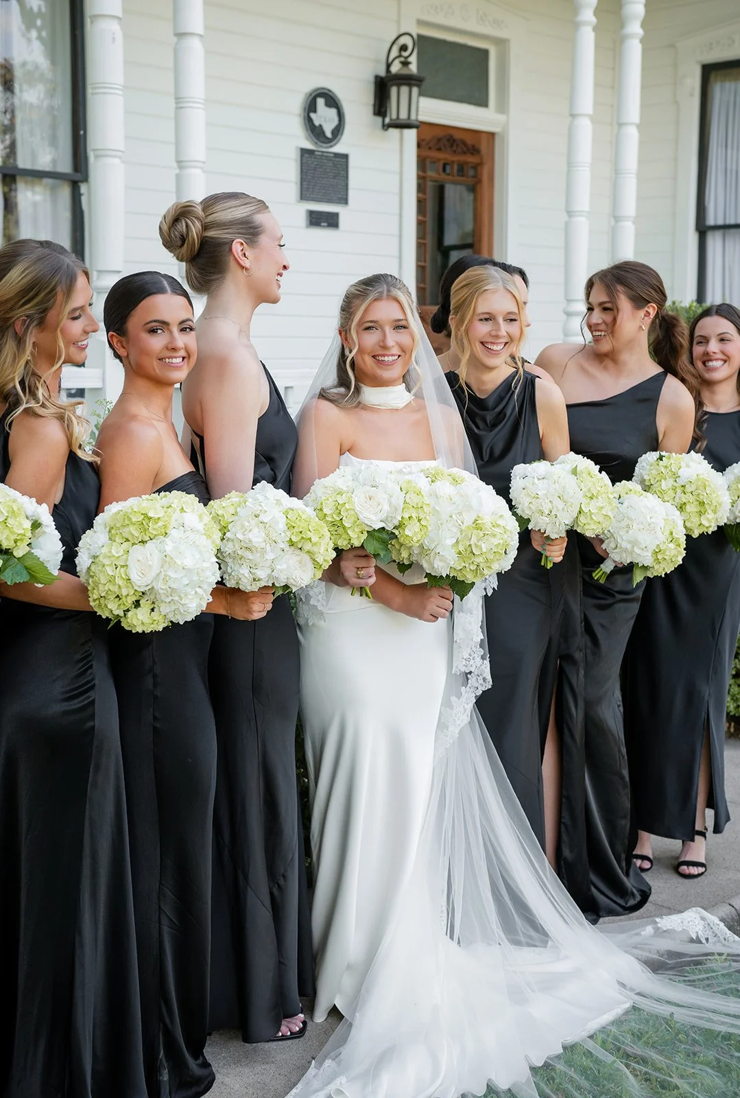 A bride standing with her bridesmaids outside a white house, all holding bouquets of white and green flowers, smiling and celebrating.