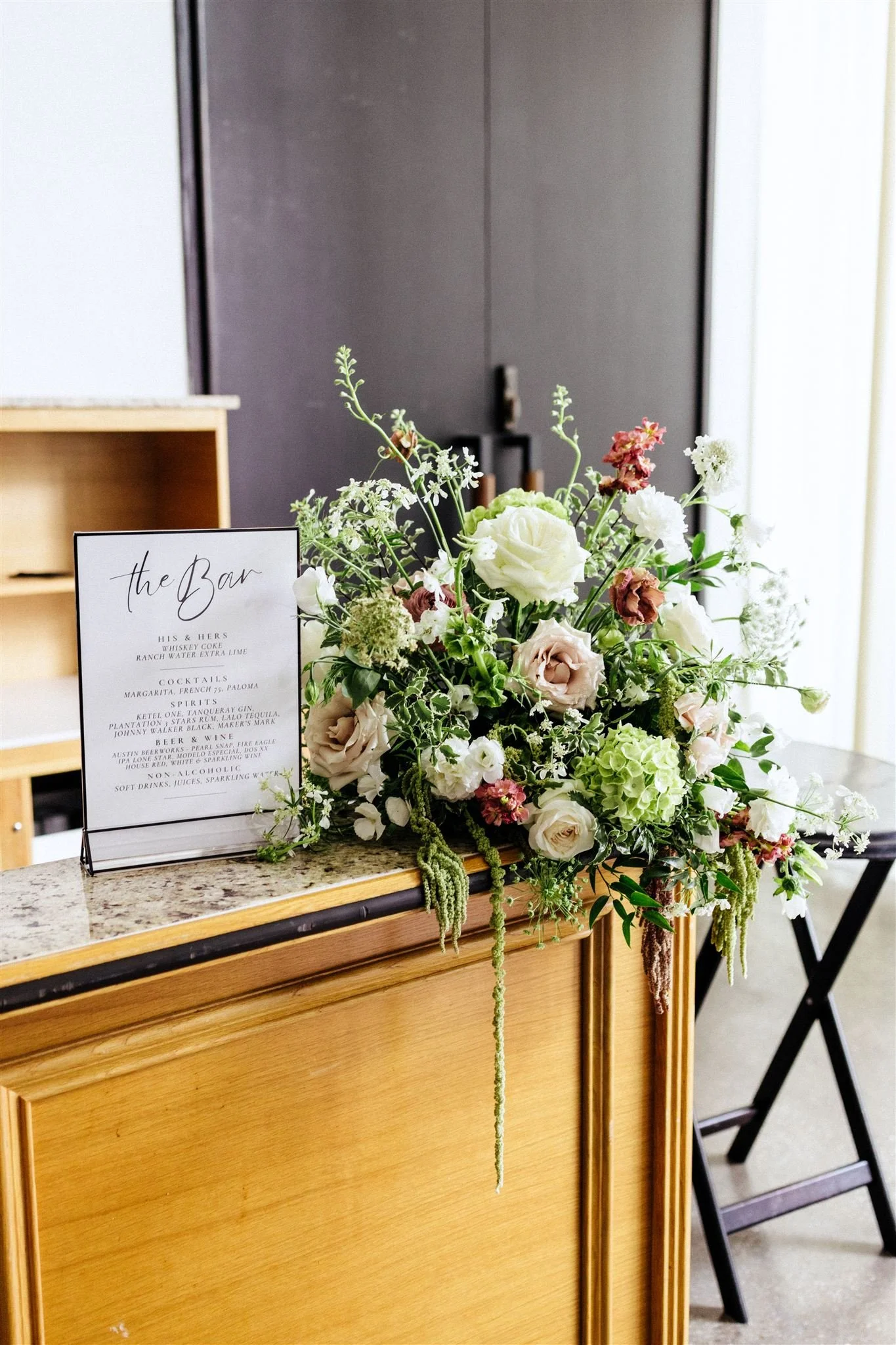 A floral centerpiece with white, pink, and green flowers on a wooden counter, with a sign that reads 'the Bar' listing drink options, in an indoor setting.