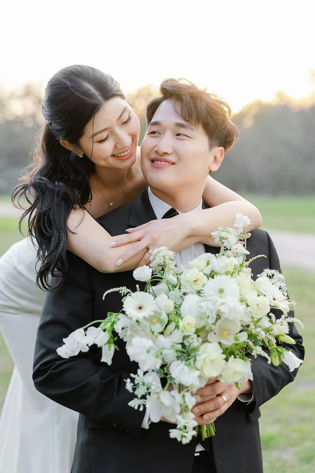 A bride and groom on their wedding day, with the bride hugging the groom from behind and both smiling. The groom is holding a large bouquet of white flowers.