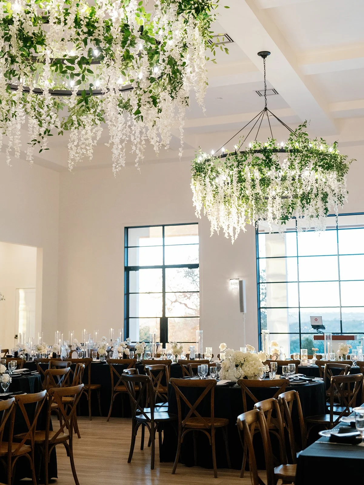 Elegant event space with large hanging floral chandeliers, round tables with white floral centerpieces, and black tablecloths, illuminated by natural light from large windows.