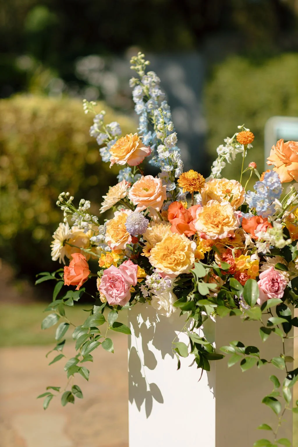 A colorful flower arrangement with pink, orange, yellow, and white flowers in a white vase outdoors.