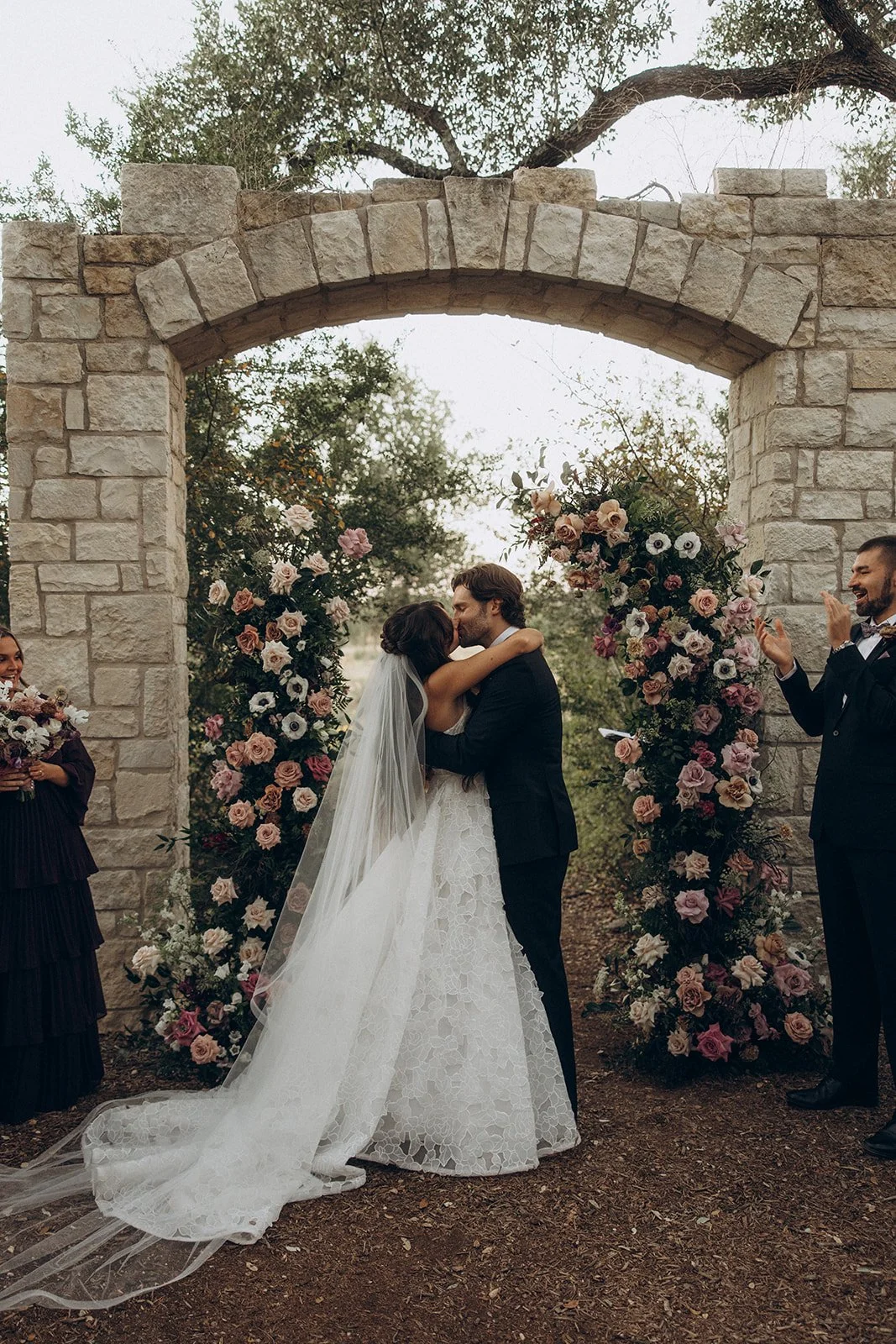 A bride and groom kiss under a stone arch surrounded by floral arrangements, with wedding guests celebrating nearby.