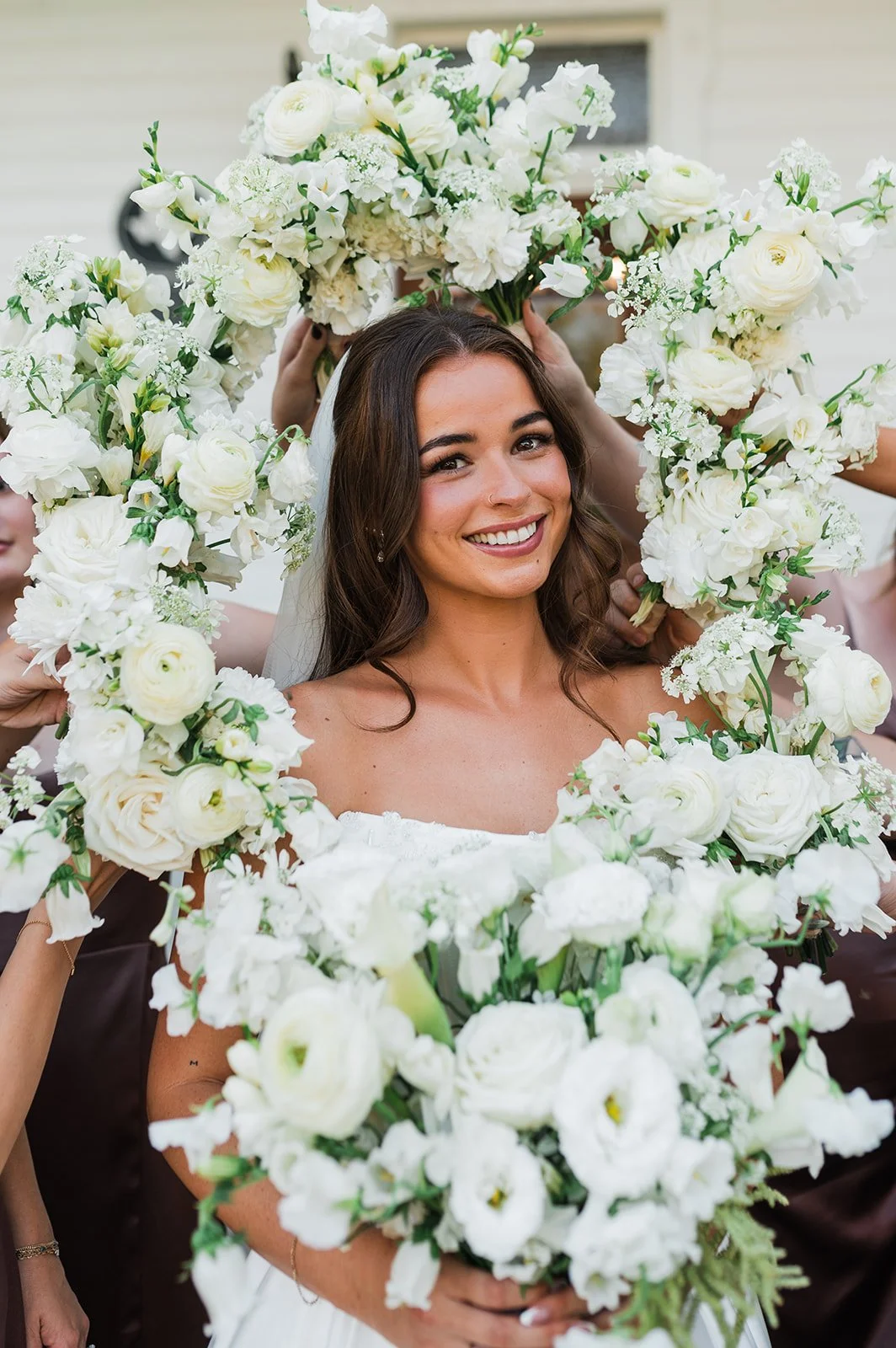 A woman with brown hair and a white dress smiling, holding a large floral arrangement of white flowers around her head.