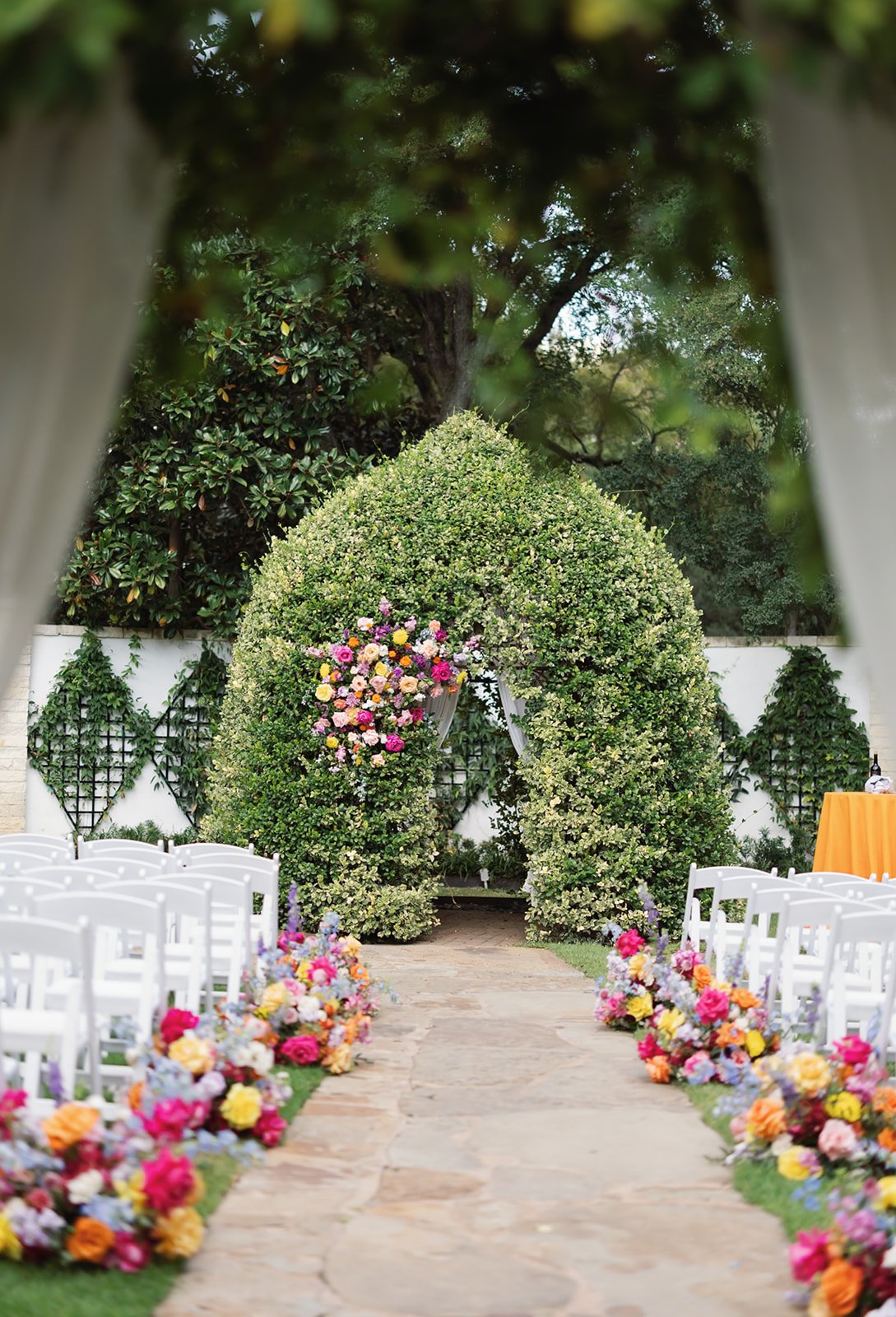 Outdoor wedding arch decorated with flowers, with white chairs lining a stone aisle, and floral arrangements on either side, set in a garden with trees and greenery.