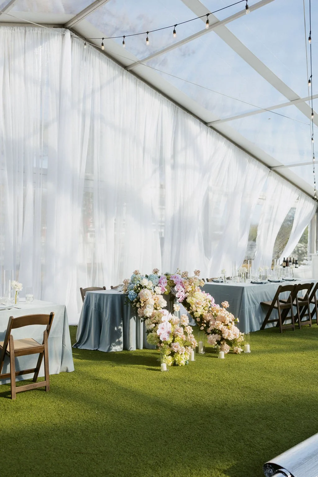 Elegant outdoor wedding reception setup inside a white tent with flowing curtains and string lights. Tables are covered with light gray tablecloths, surrounded by wooden chairs, and decorated with a large bouquet of pink and white flowers. Candles ar