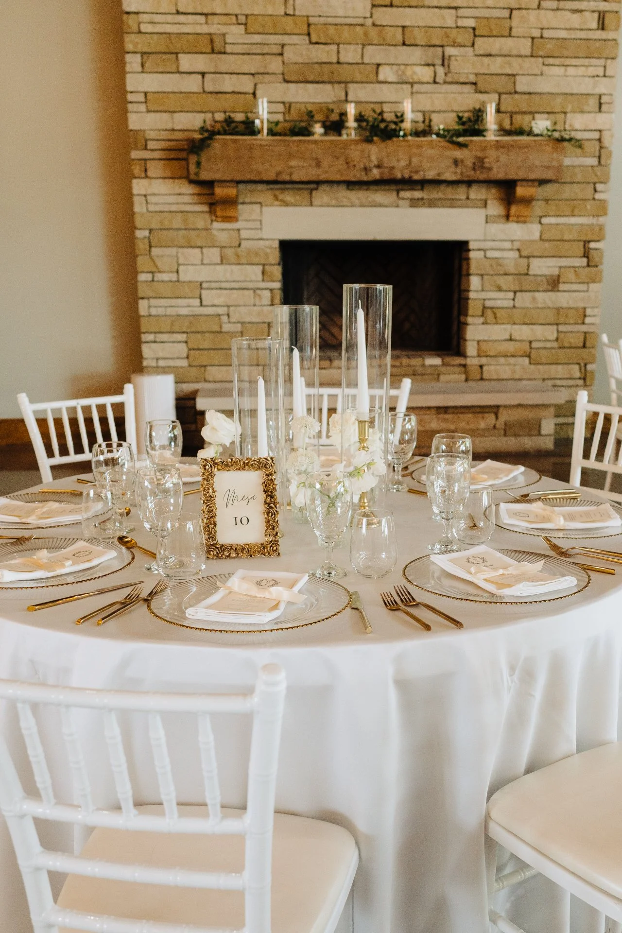 A round table set for a formal event with white tablecloth, gold flatware, wine glasses, white napkins, and a decorative centerpiece with candles and flowers. A small framed sign with the word 'Mesa' and the number '10' is on the table. The backgroun