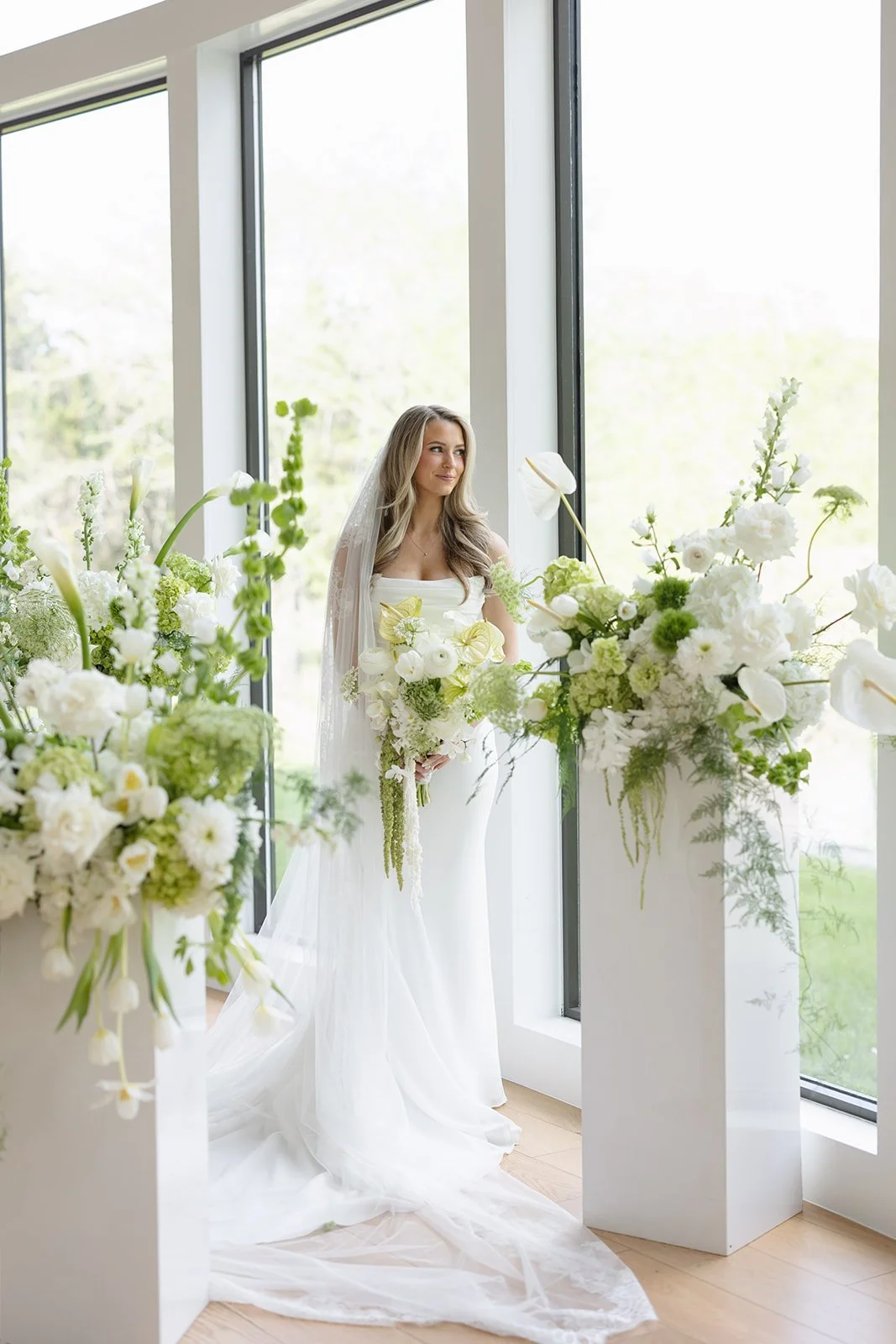 A bride in a white wedding dress standing next to large white and green floral arrangements near floor-to-ceiling windows.
